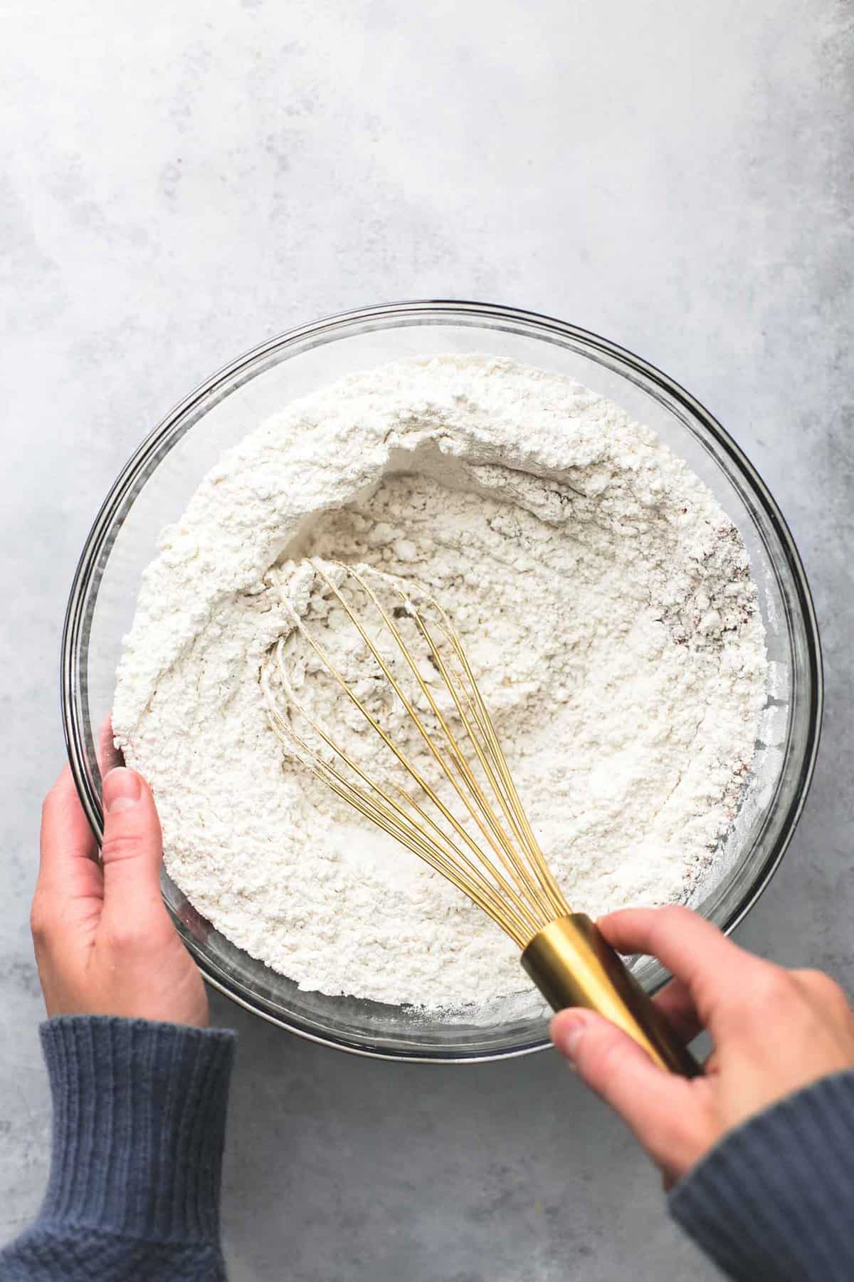 top view of hands holding a glass bowl of flour mixture with the other hand holding a whisk in the bowl.