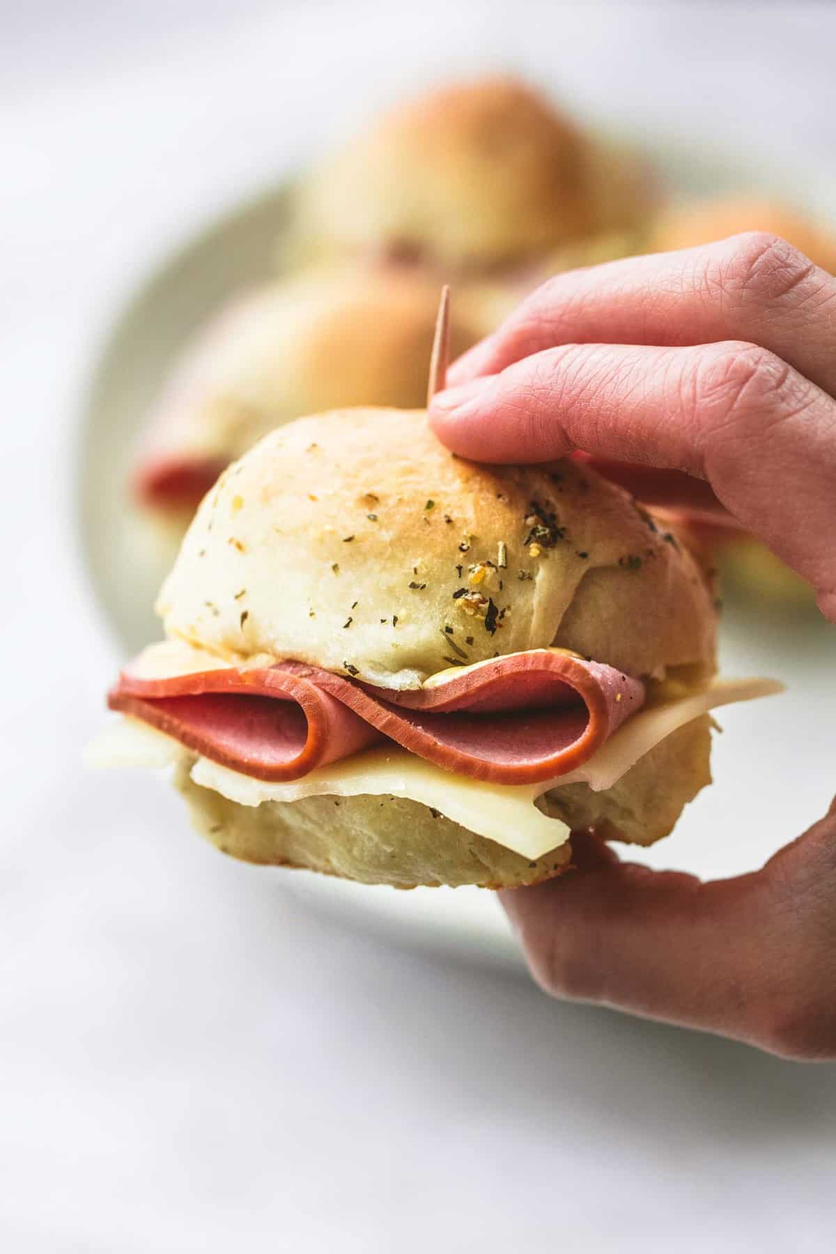 a hand holding a ham and cheese slider on a buttery herb roll with more sliders on a plate faded in the background.