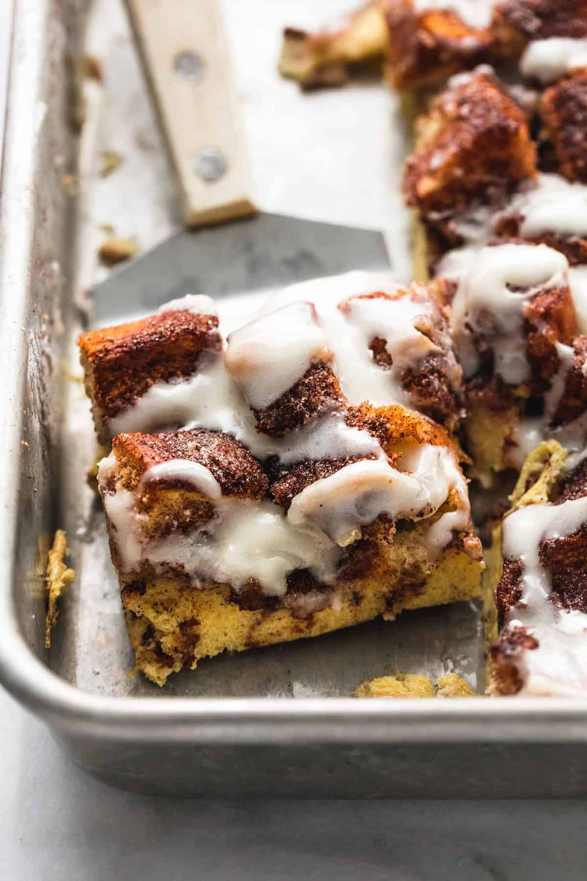 close up of a piece of overnight cinnamon roll French toast casserole on a spatula next to the rest of the casserole on a baking sheet.