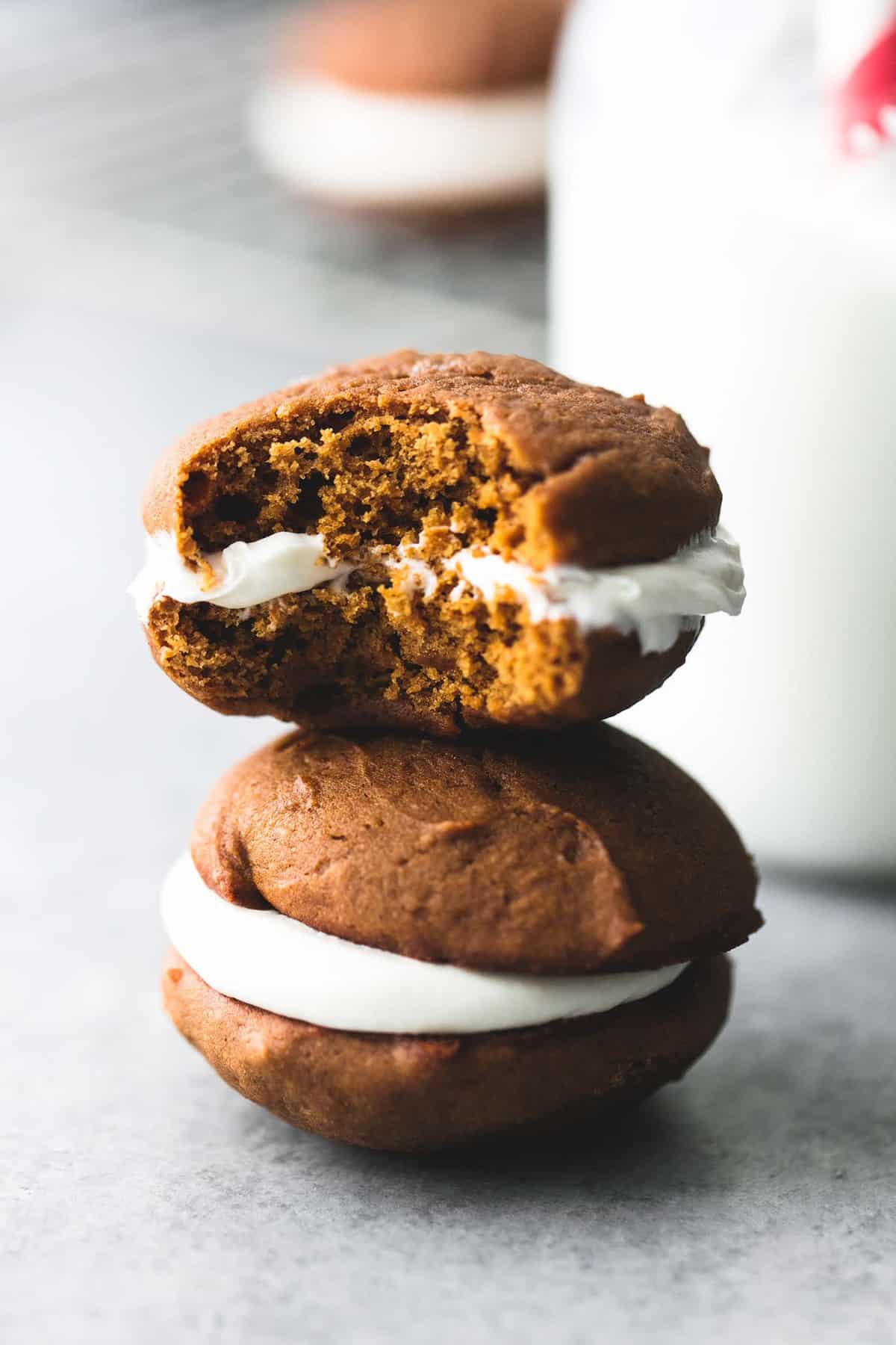 stacked gingerbread whoopie pies with the top one missing a bite with a jar of milk with a straw and more pies on a cooling rack faded in the background.