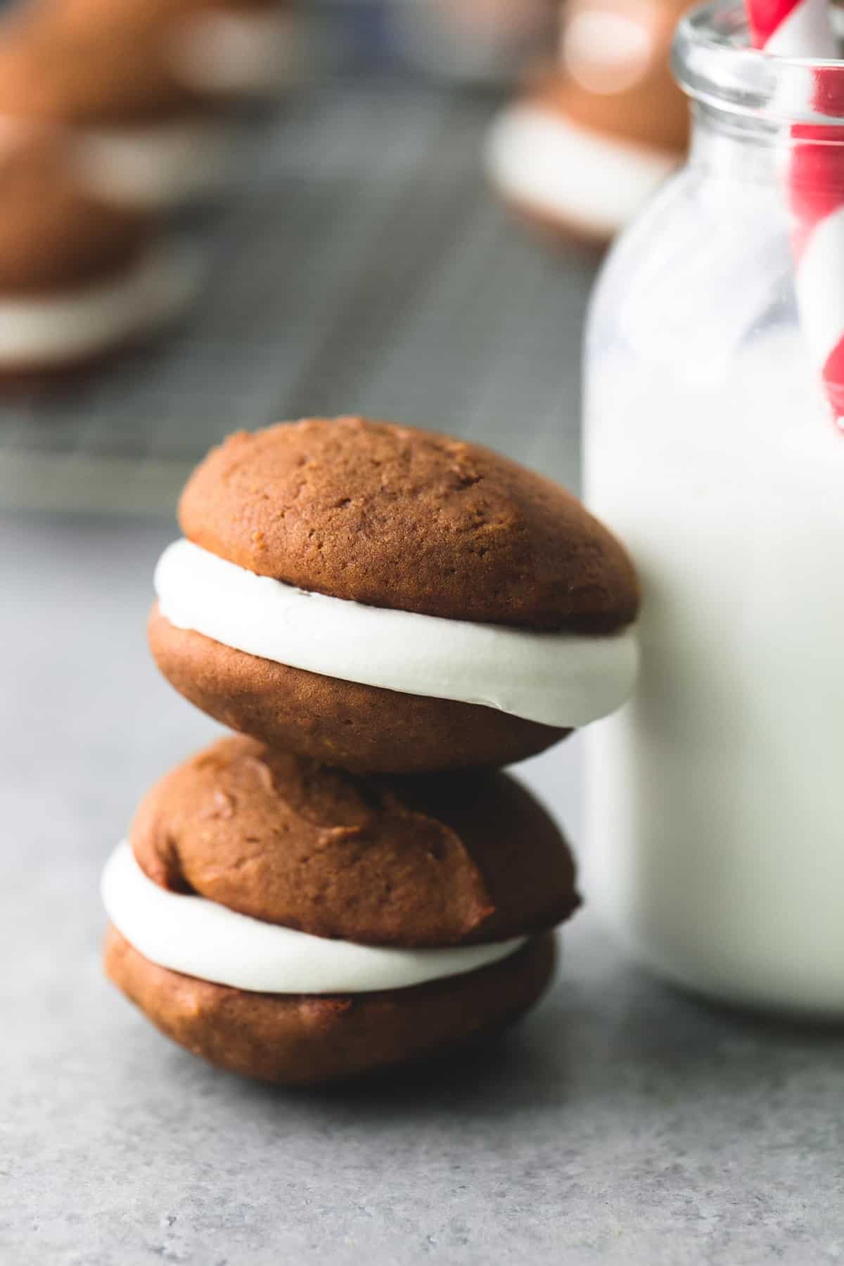 stacked gingerbread whoopie pies leaning against a jar of milk with a straw with more pies on a cooling rack faded in the background.