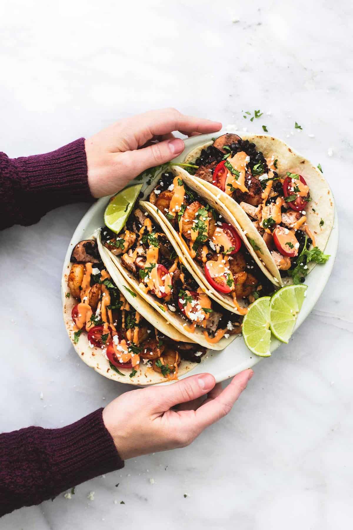 a pair of hands holding a plate of shrimp or chicken and lentil tacos with lime slices on the side.