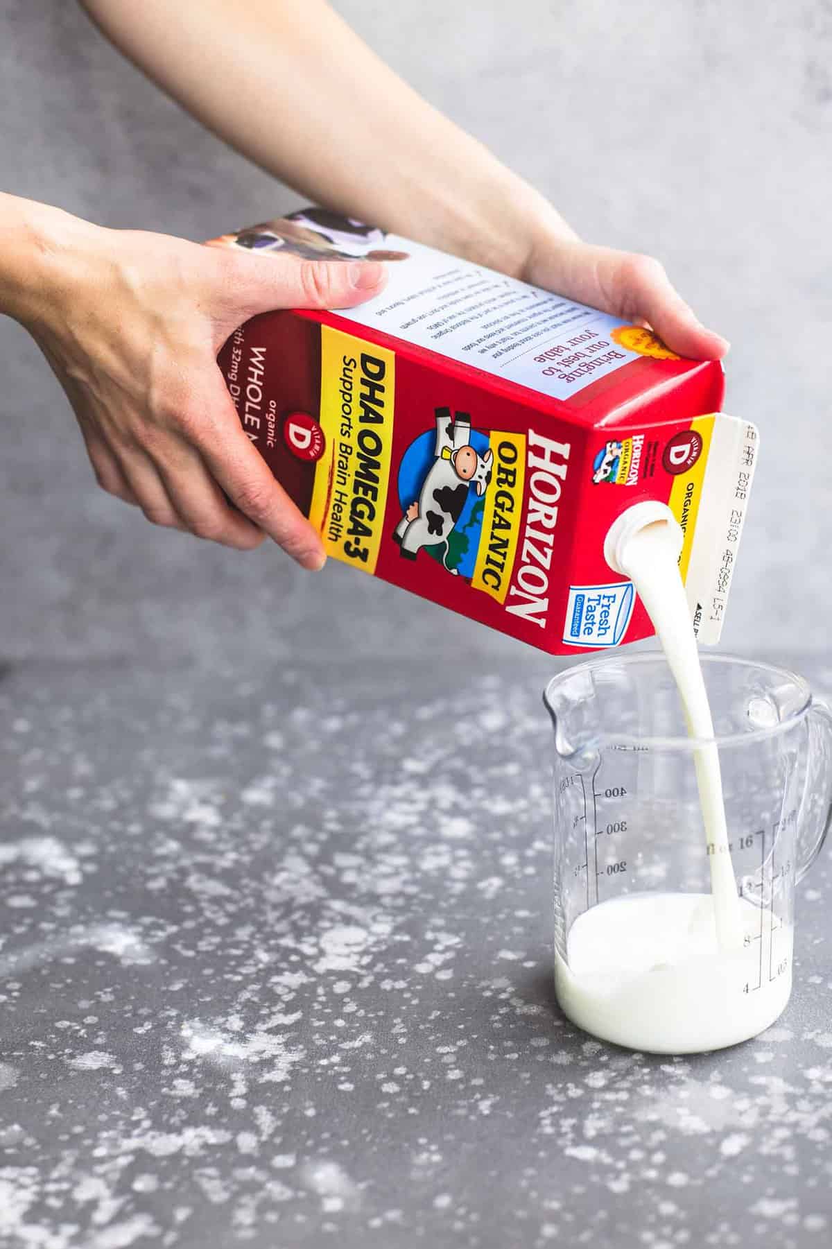 a pair of hands pouring a carton of Horizon Organic whole milk into a liquid measuring cup.