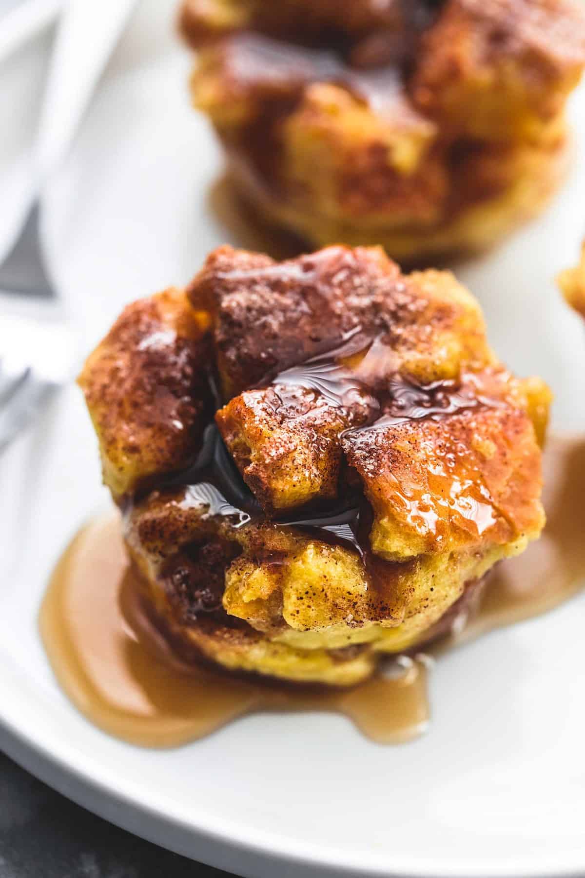 close up of a baked cinnamon French toast muffin topped with syrup with a fork and more muffins in the background all on a plate.