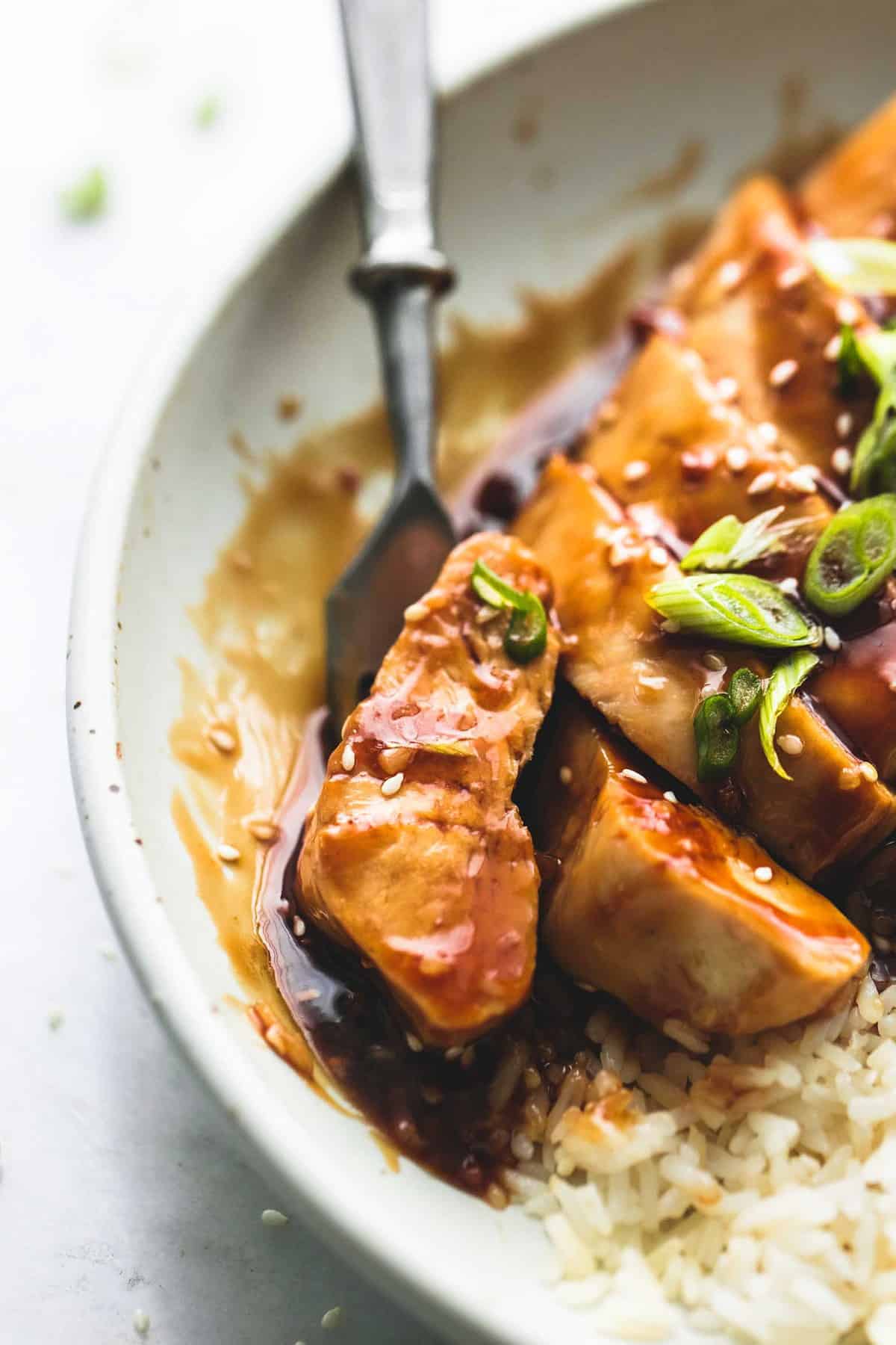 close up of a piece of baked teriyaki chicken on a fork in front of the rest of the chicken and rice all on a plate.