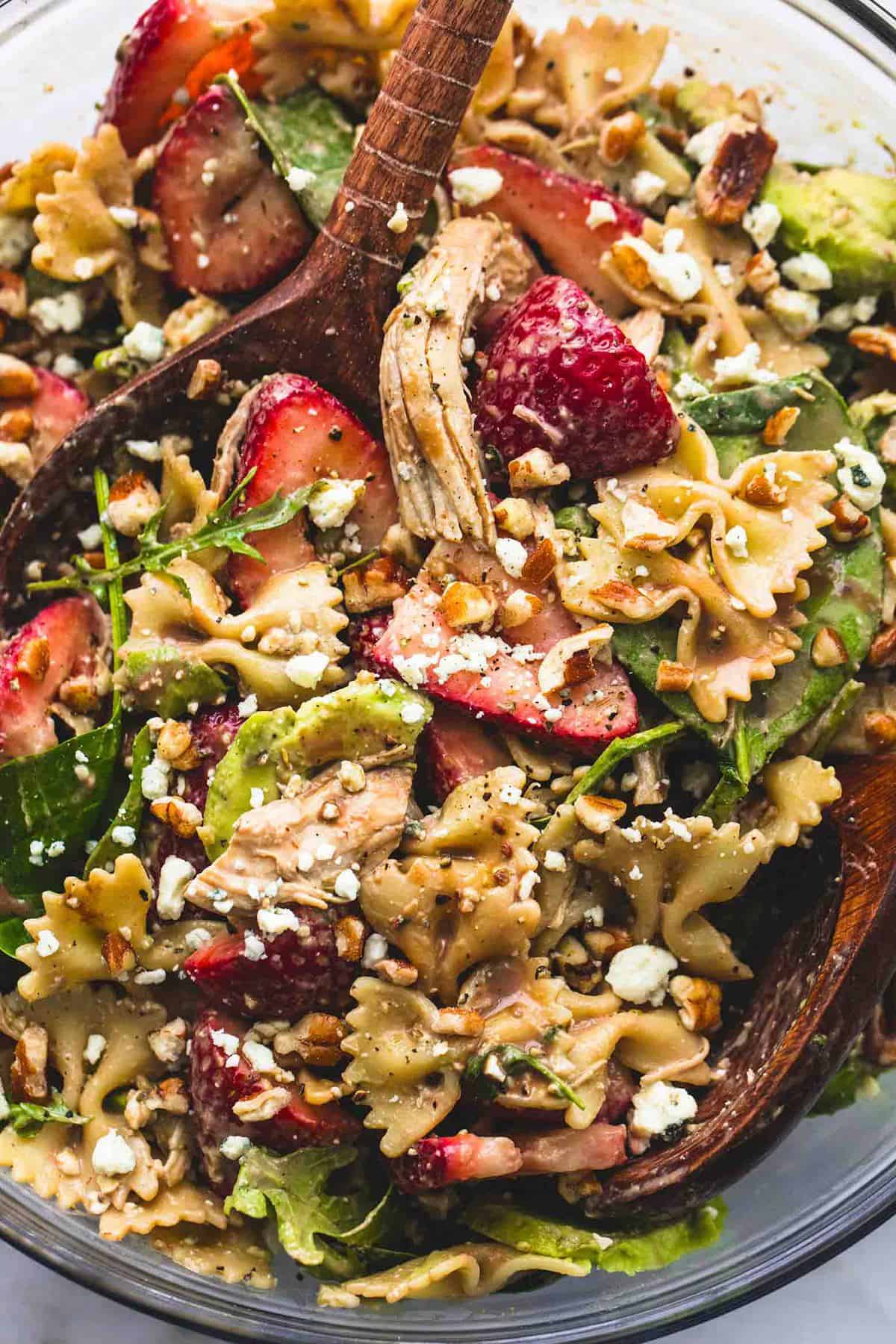 close up top view of strawberry avocado chicken pasta salad with two wooden serving spoons in a glass bowl.