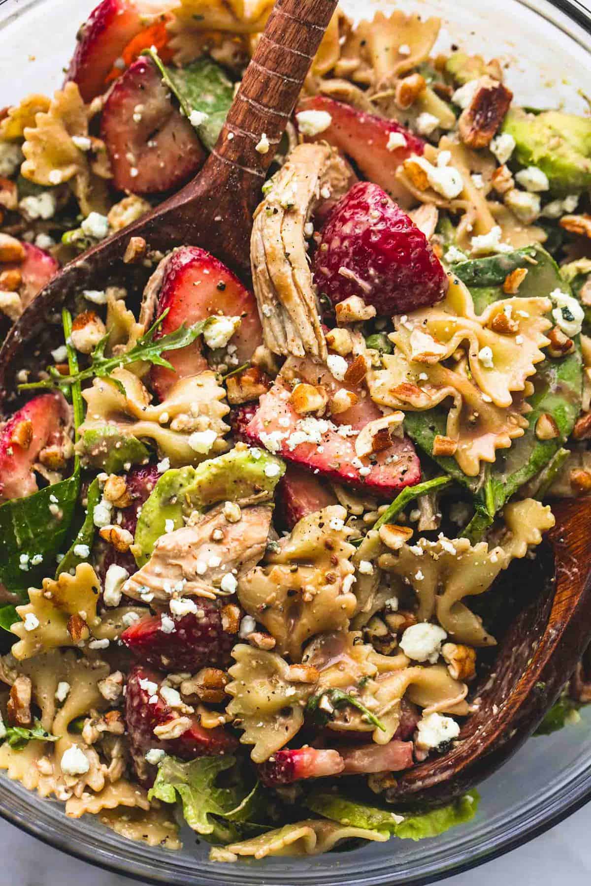 Strawberry Avocado Chicken Pasta Salad | lecremedelacrumb.com close up top view of strawberry avocado chicken pasta salad with two wooden serving spoons in a glass bowl.