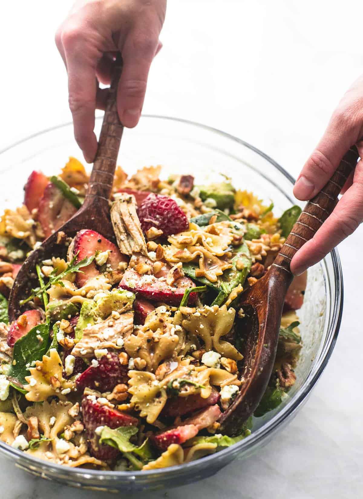 a pair of hands scooping some strawberry avocado chicken pasta salad from a glass bowl with two wooden serving spoons.