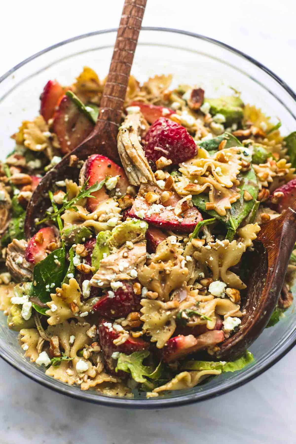 top view of strawberry avocado chicken pasta salad with two wooden serving spoons in a glass bowl.