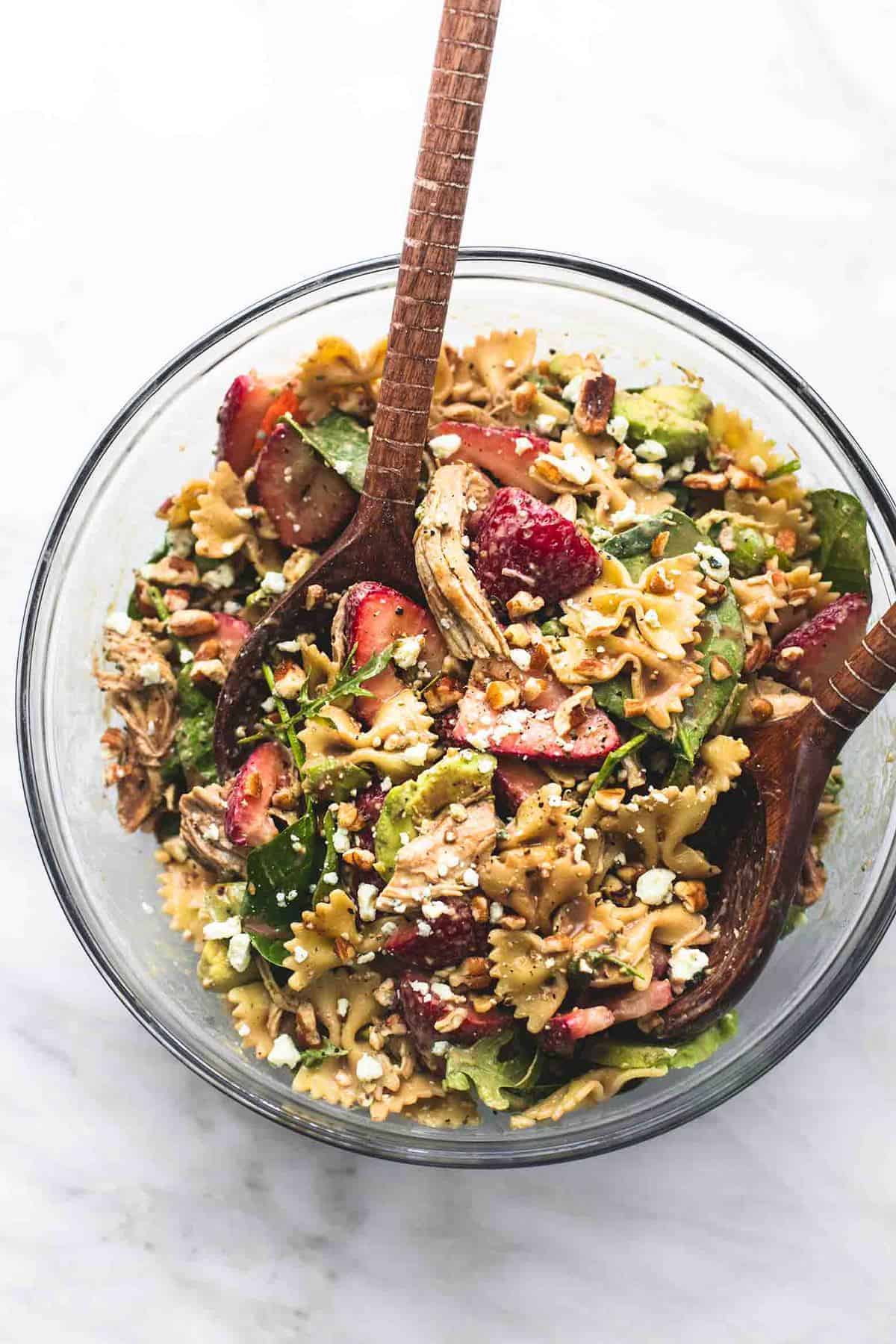 top view of strawberry avocado chicken pasta salad with two wooden serving spoons in a glass bowl.