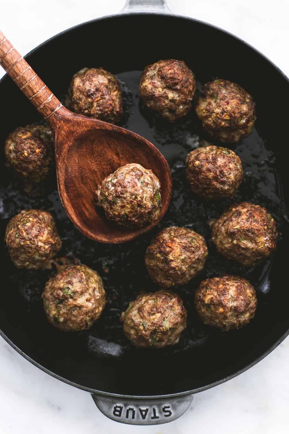 top view of a Greek meatball sitting on top of a wooden serving spoon above a pan of more meatballs.