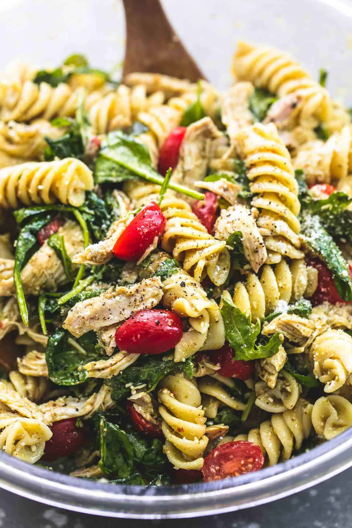 close up of pesto chicken pasta salad with a wooden serving spoon in a glass bowl.