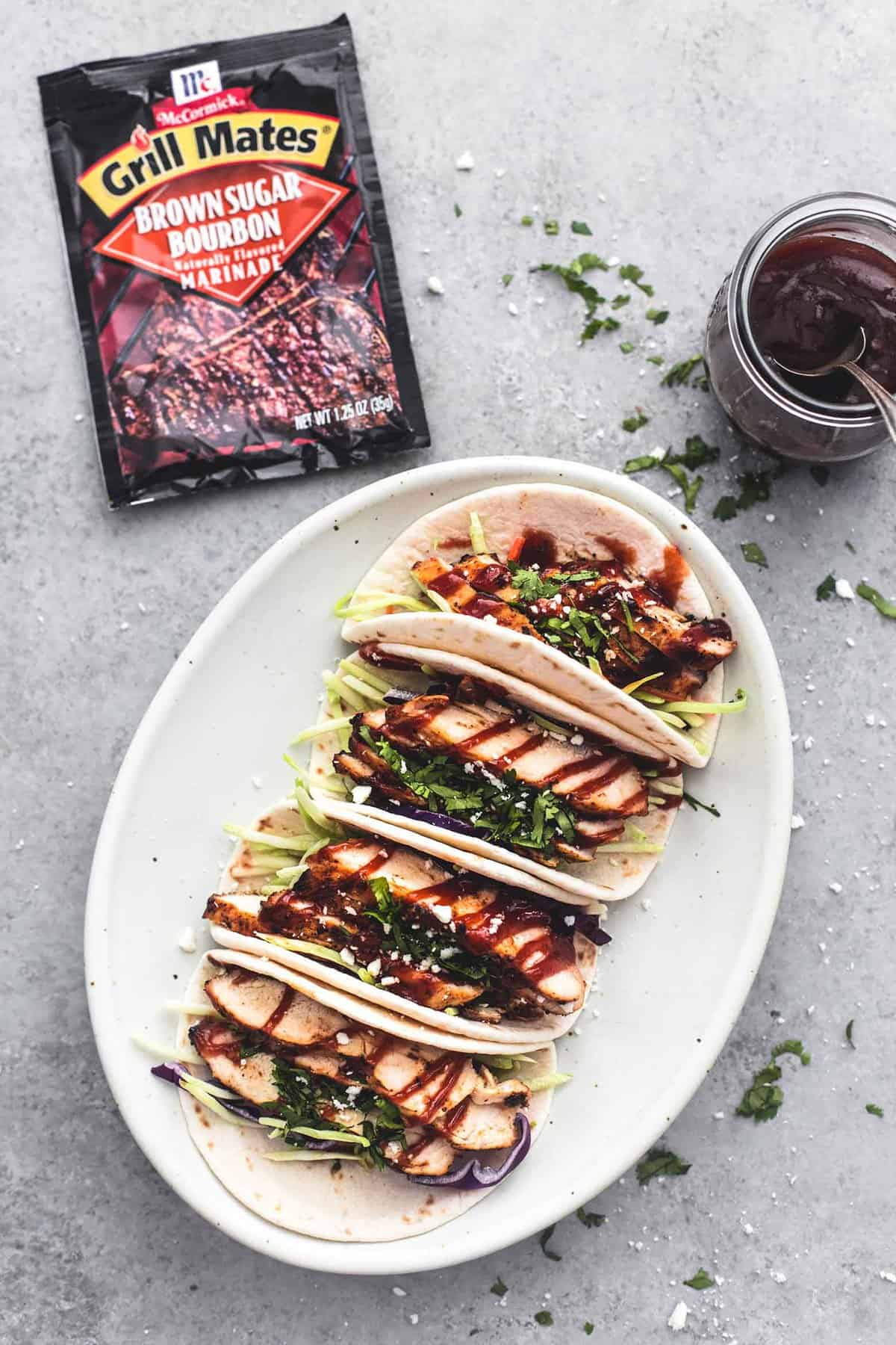 top view of mini grilled bbq chicken tacos on a plate with bbq sauce with a spoon in a jar and a package of Grill Mates on the side.