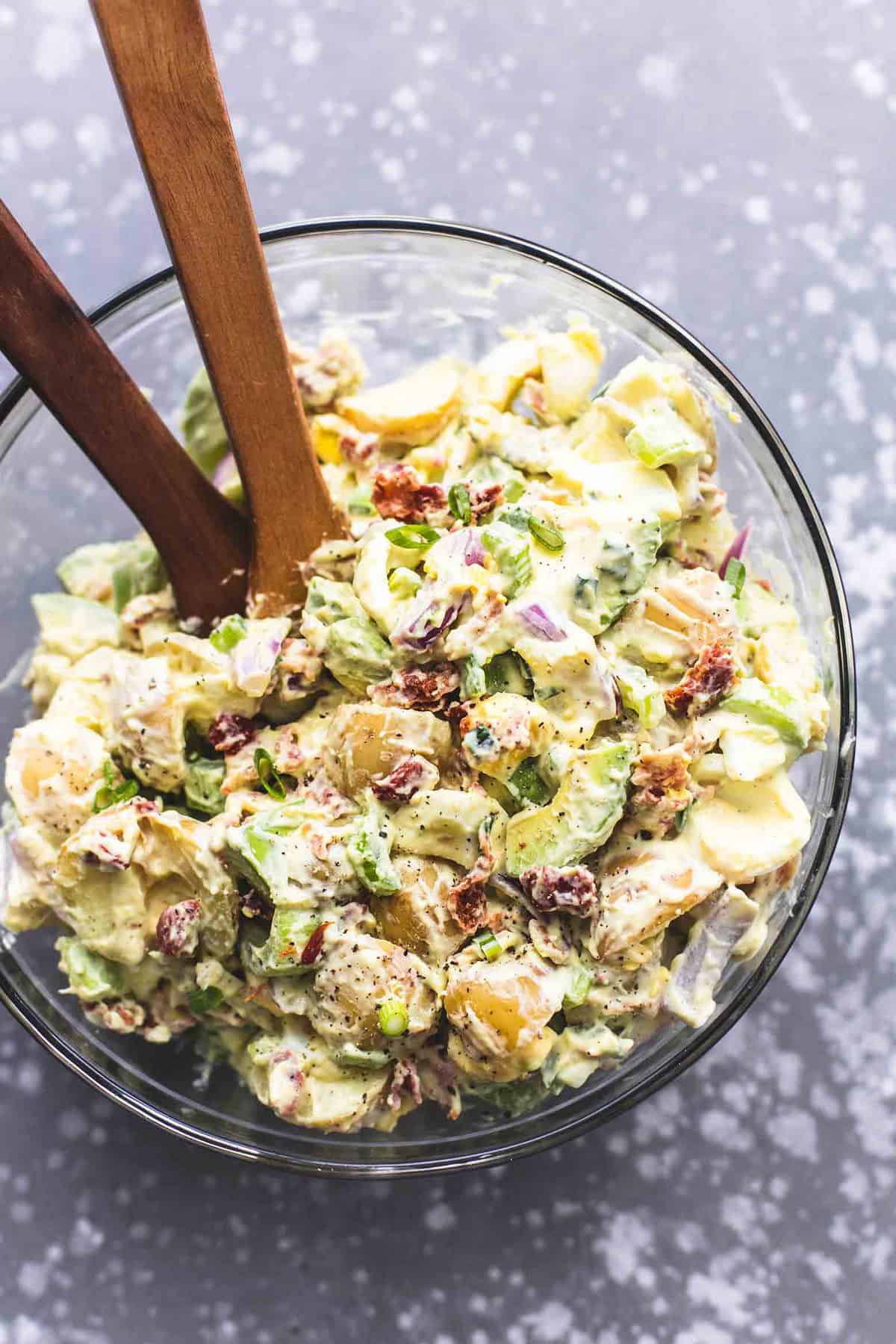 top view of bacon avocado potato salad with two wooden serving spoons in a glass bowl.