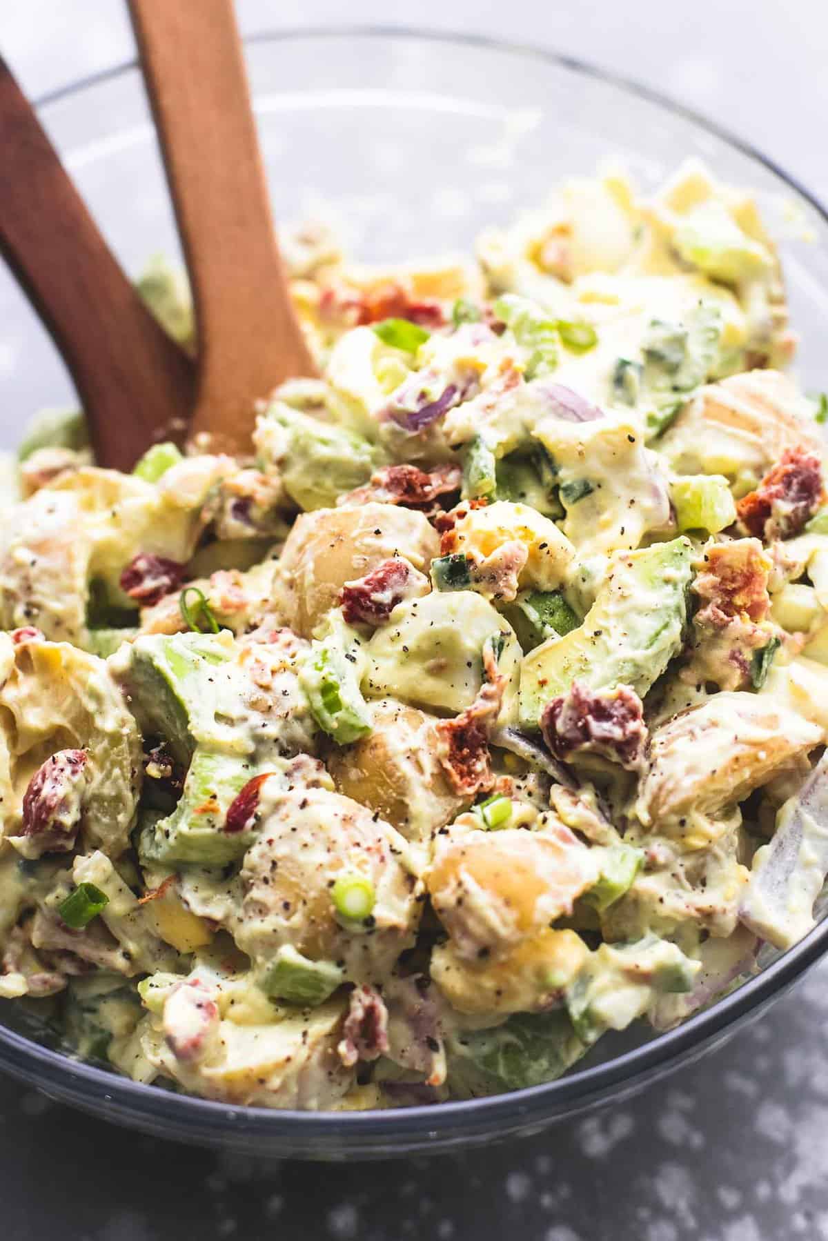 bacon avocado potato salad with two wooden serving spoons in a glass bowl.