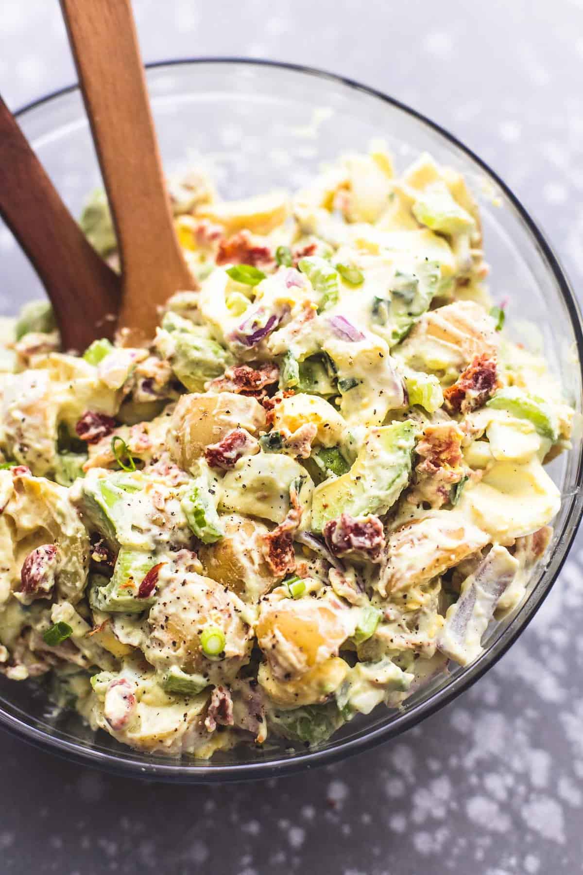 top view of bacon avocado potato salad with two wooden serving spoons in a glass bowl.