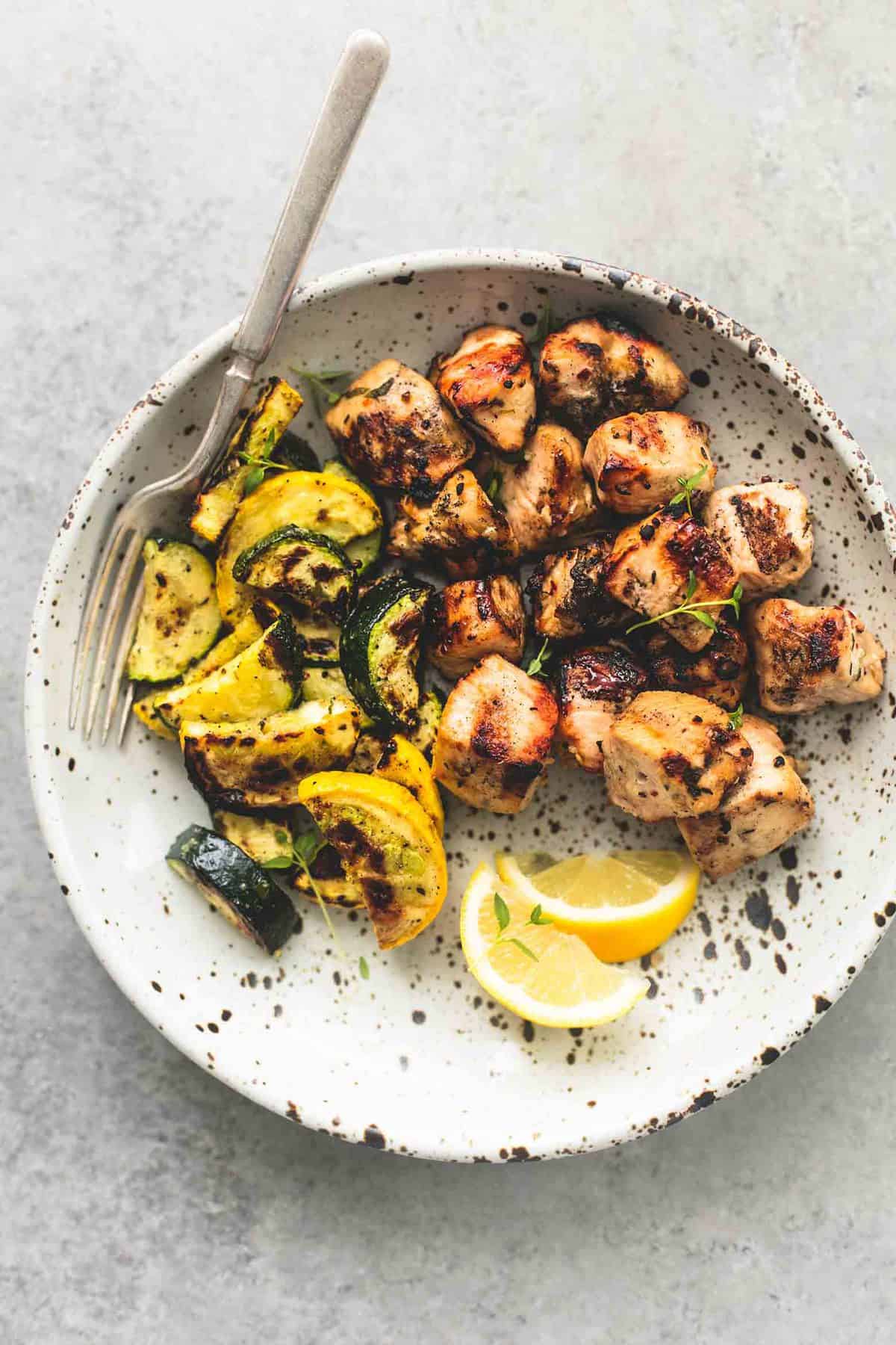 top view of deconstructed herb chicken and veggie skewers with lemon slices and a fork on a plate.