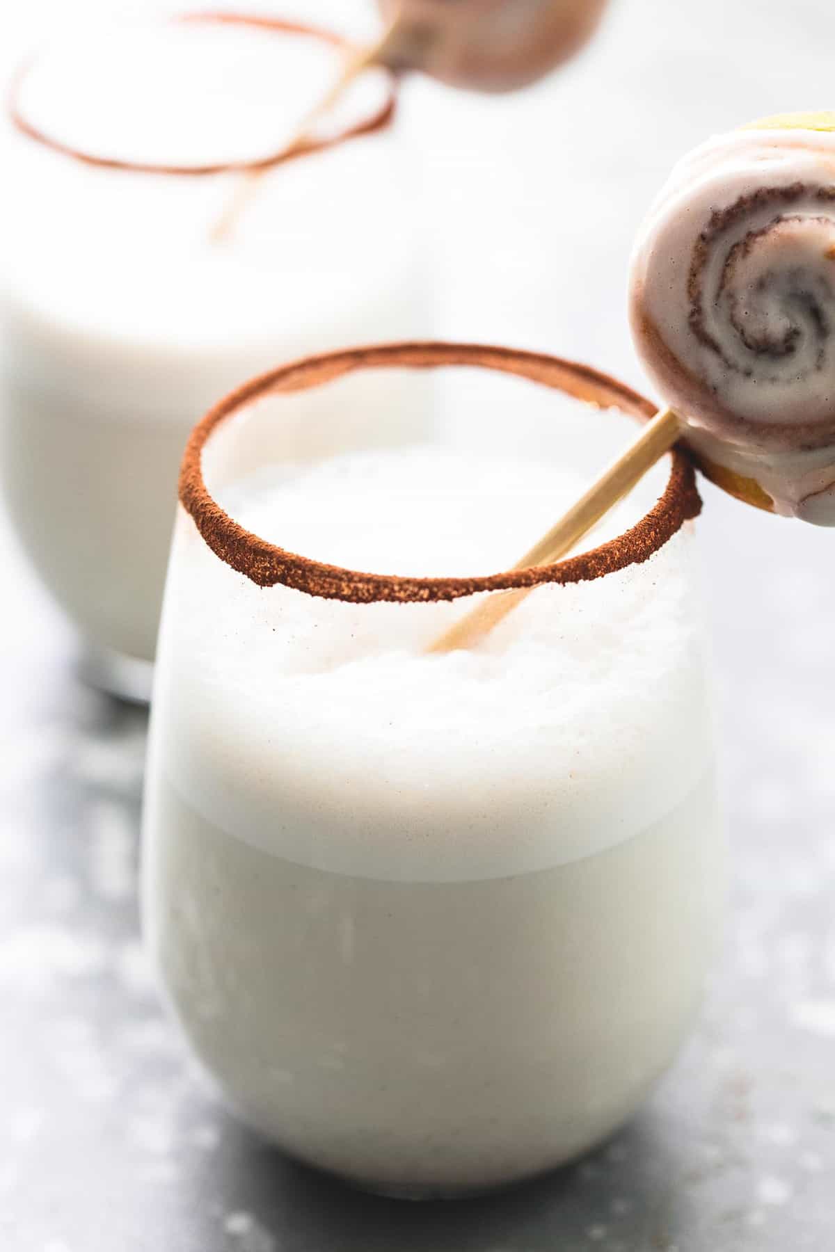 a glass of cinnamon bun moon milk and a mini cinnamon roll on a stick sitting in it with cinnamon around the ring of the cup with a mother glass in the background.