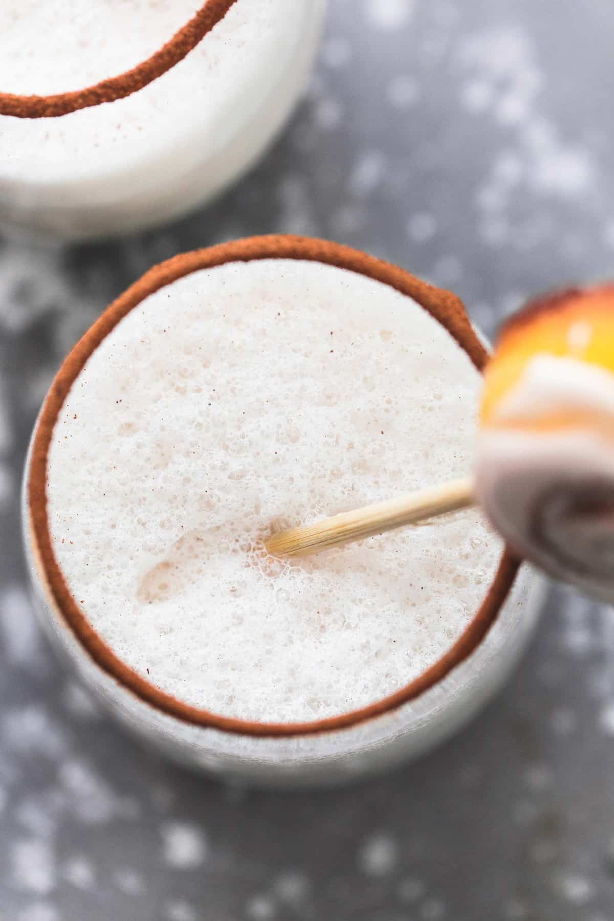 top view of cinnamon bun moon milk in a glass with a mini cinnamon roll on a stick in it and cinnamon around the ring of the glass with another glass of milk on the side.