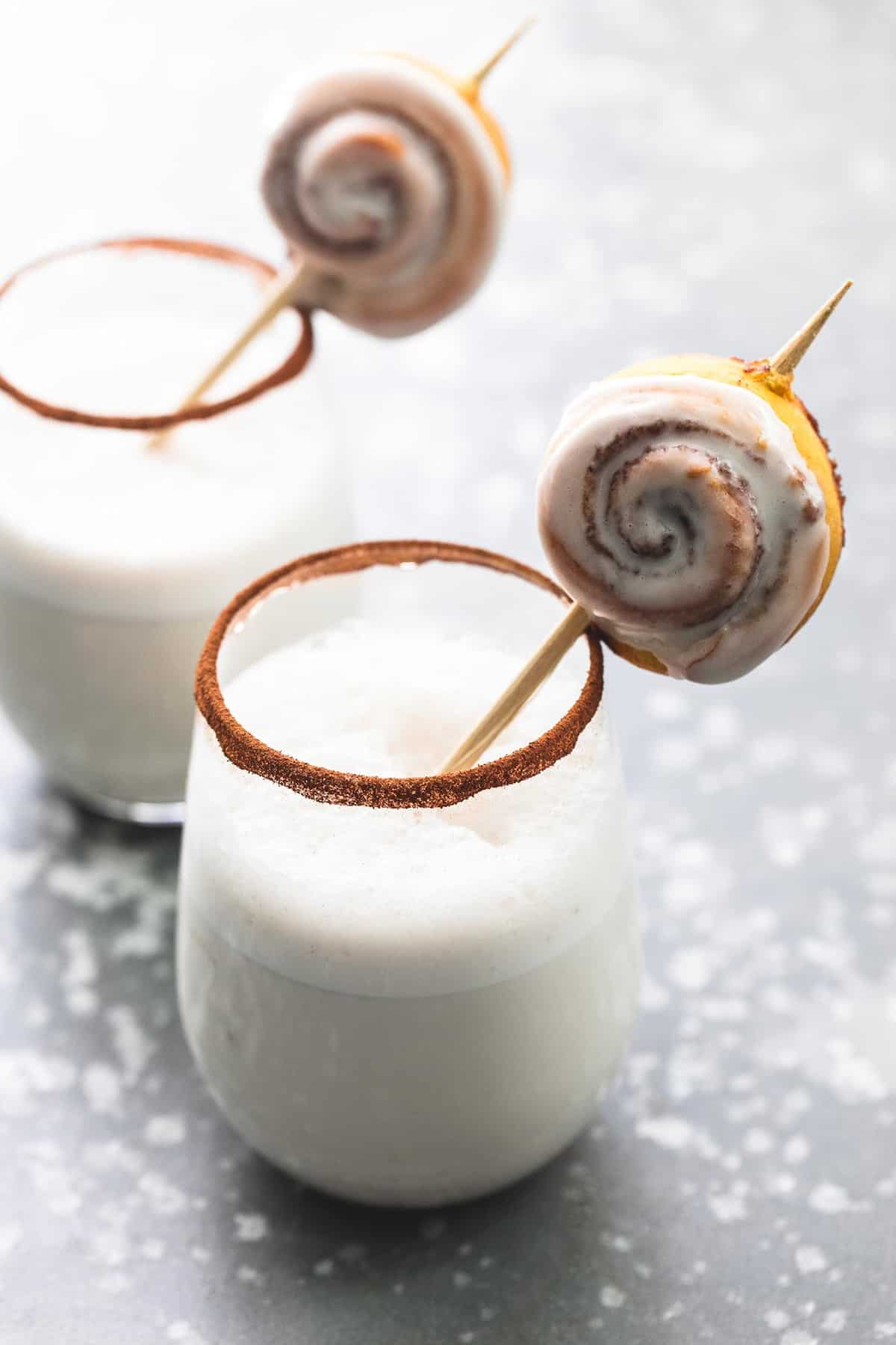 glasses of cinnamon bun moon milk with a mini cinnamon roll on a stick sitting in it with cinnamon around the ring of the glasses.