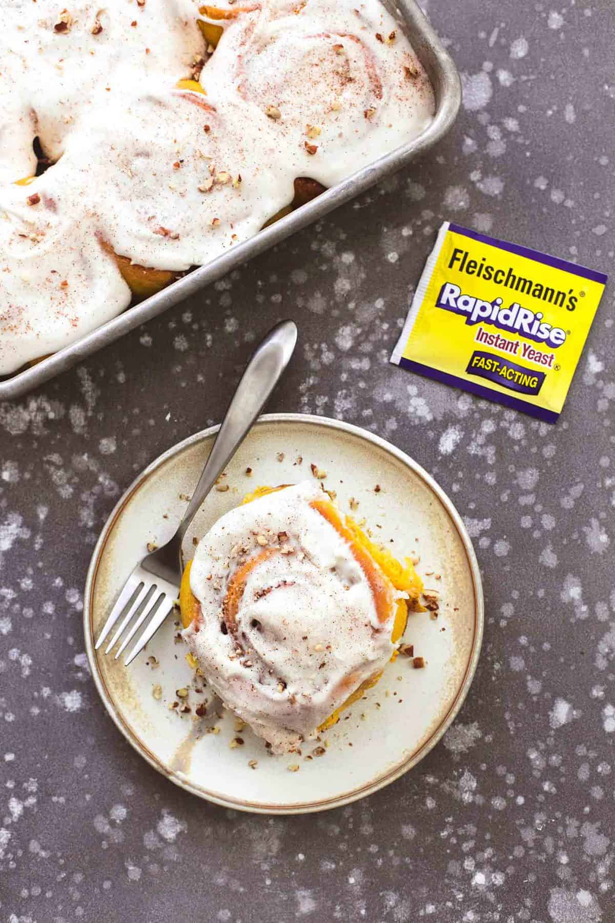 top view of a pumpkin cinnamon roll with a fork on a plate with a packet of RapidRise yeast and a baking sheet of more cinnamon rolls on the side.