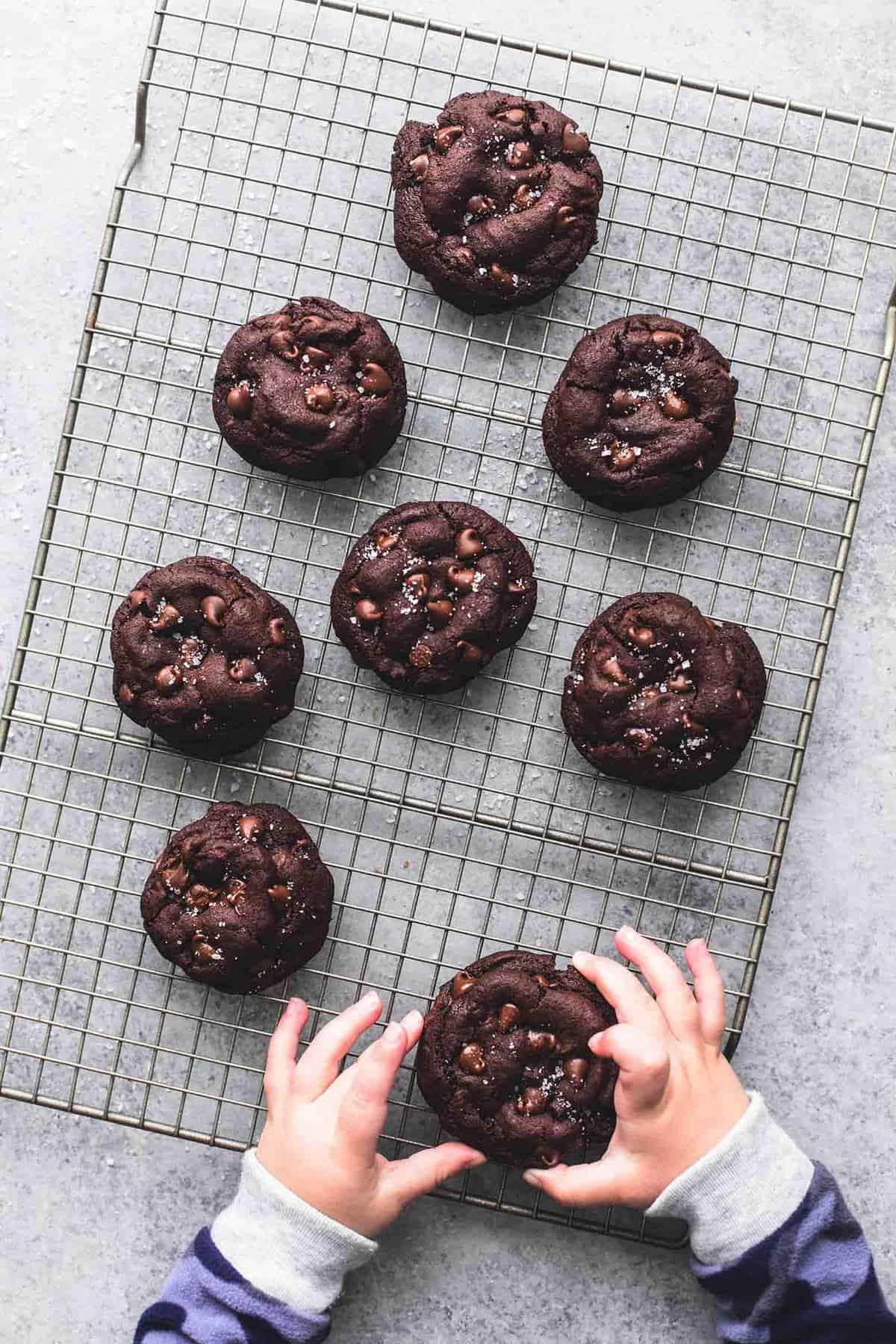 top view of salted caramel stuffed double chocolate cookies on a cooling rack with a child's hands grabbing a cookie.