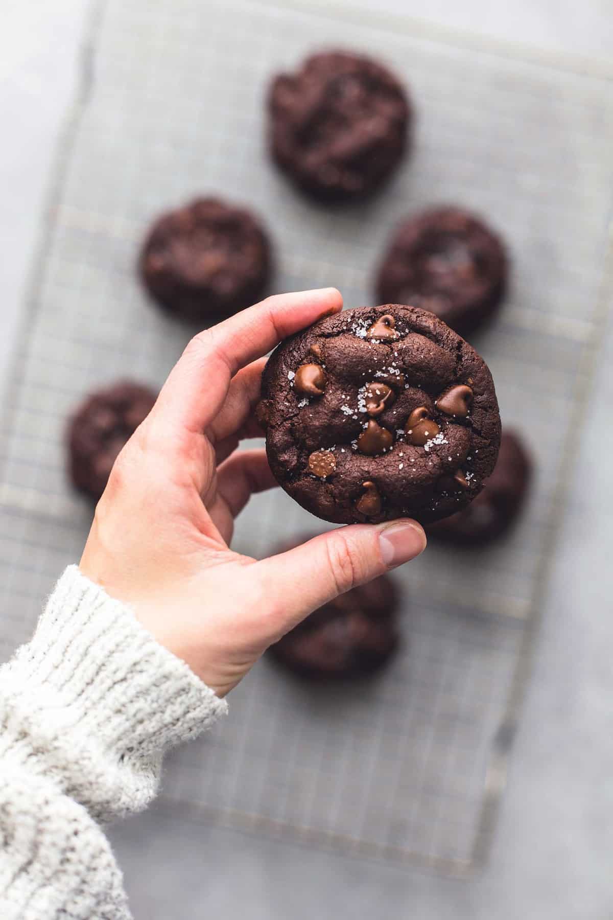 a hand holding a salted caramel stuffed double chocolate cookie above a cooling rack with more cookies.