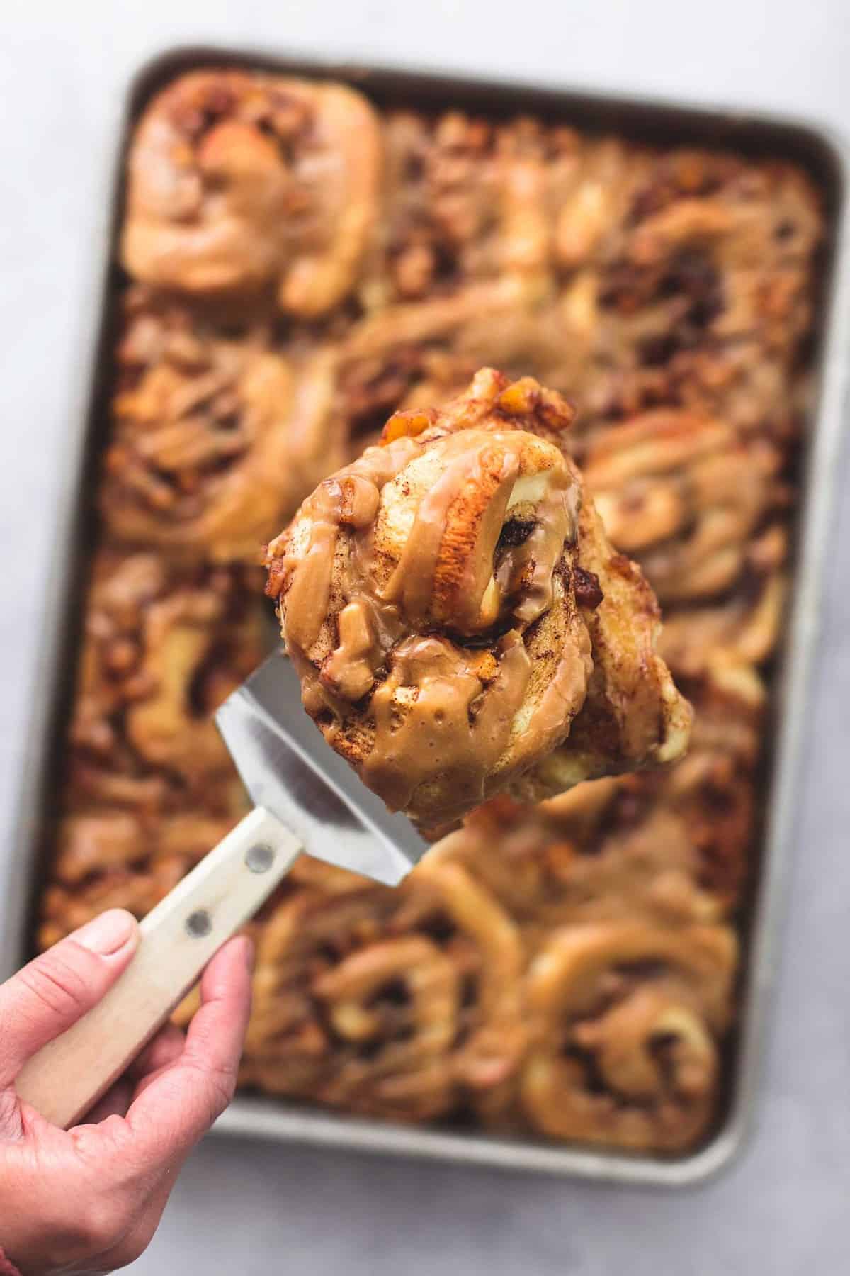a hand holding up a spatula with a apple pie cinnamon roll on it above a baking sheet of rolls.