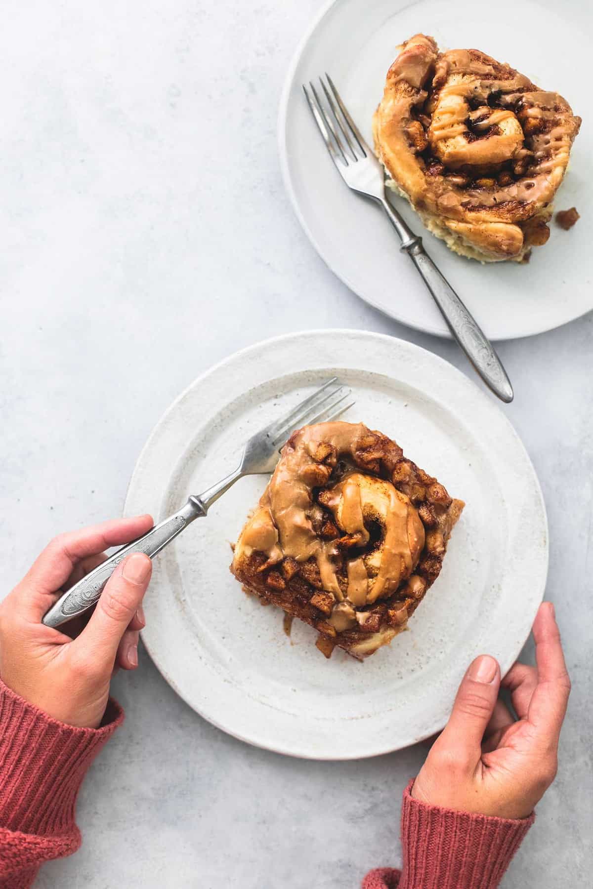top view of a pair of hands holding a plate with a apple pie cinnamon roll on it with one hand and a fork with the other with another roll and fork on a plate on the side.