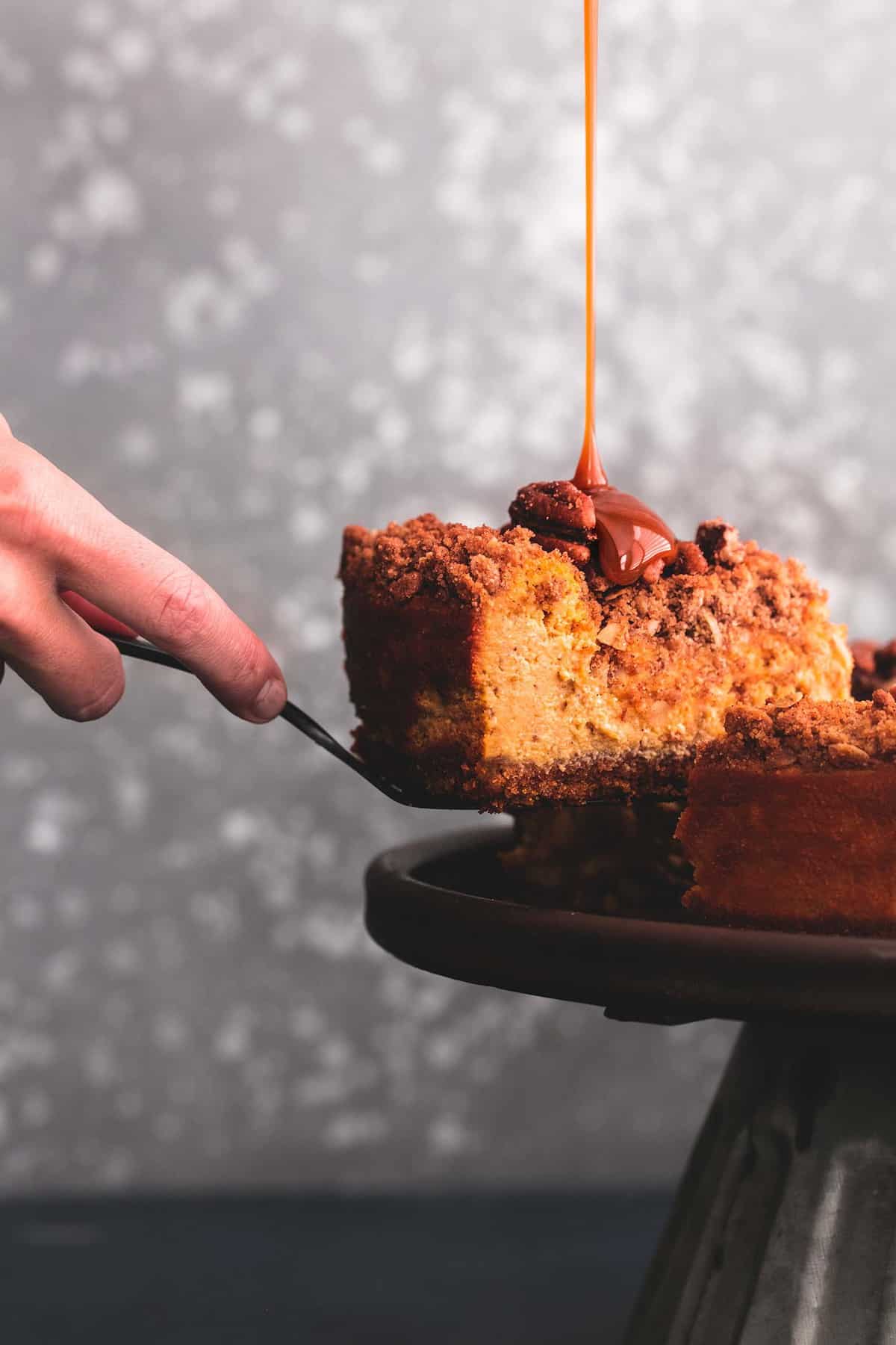 a hand separating a slice of caramel pecan pumpkin cheesecake from the rest of the cheesecake with a spatula and caramel sauce being poured on top of the slice.