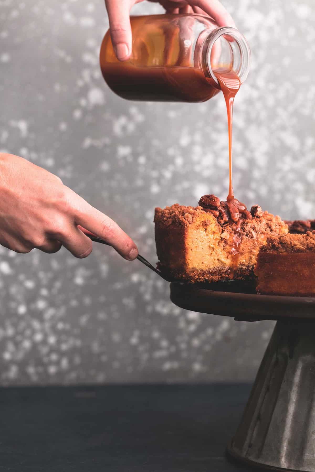 a hand separating a slice of caramel pecan pumpkin cheesecake from the rest of the cheesecake and pouring caramel sauce on the slice with the other hand.