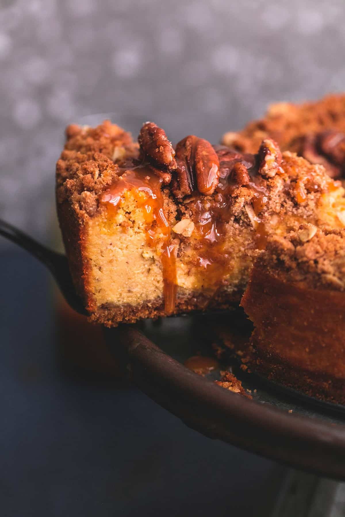 a slice of caramel pecan pumpkin cheesecake being taken from the rest of the cheesecake on a cake stand by a spatula.