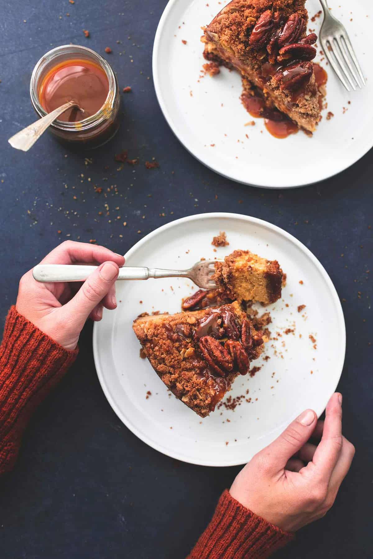 top view of a pair of hands holding a plate with a slice of caramel pecan pumpkin cheesecake on it and lifting a bite with a fork with the other hand with a jar of caramel sauce and another slice of cheesecake on a plate on the side.