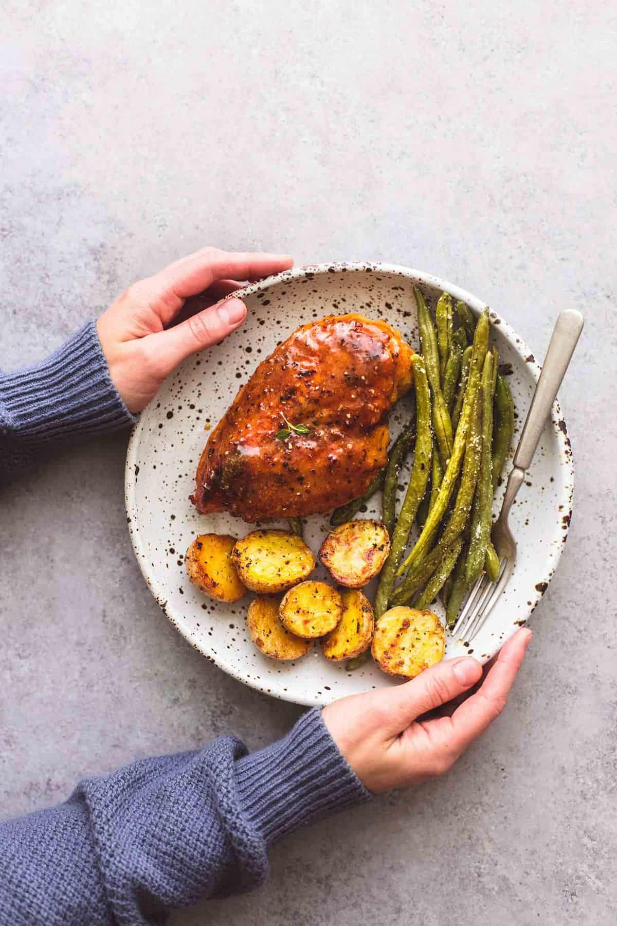 top view of a pair of hands holding a plate with sheet pan chicken potatoes and green beans and a fork on it.