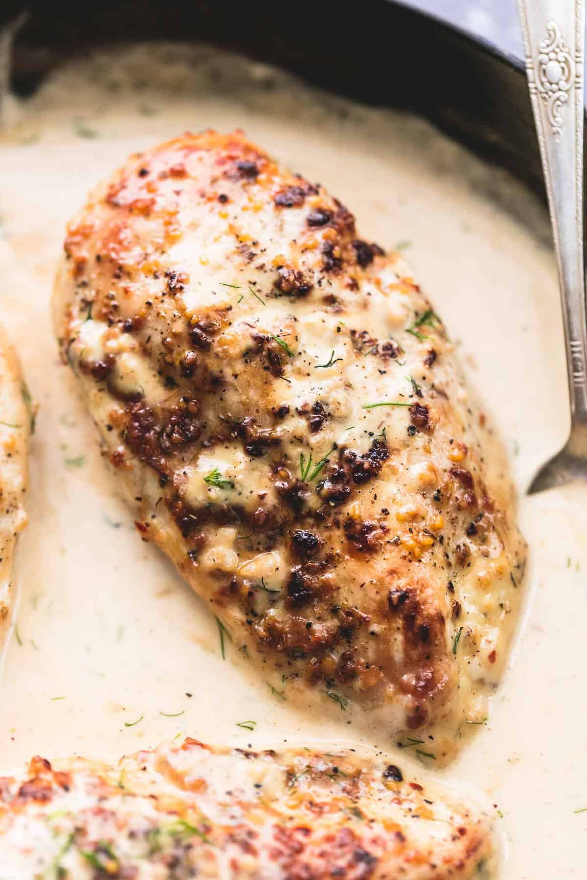 close up of a piece of lemon chicken in creamy dill sauce with more chicken and a spoon on the side all in a skillet.