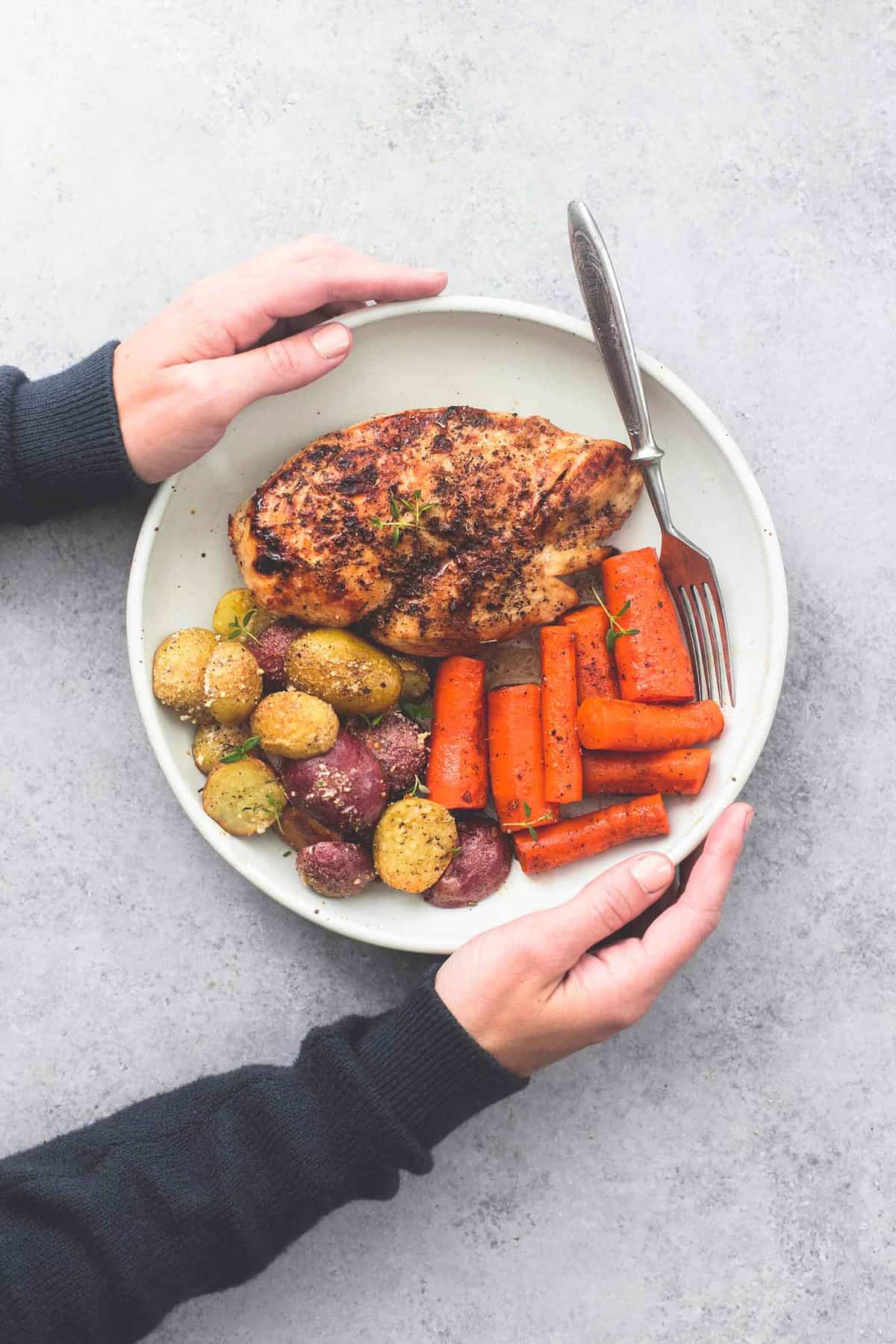 top view of a pair of hands holding a plate with a fork and sheet pan balsamic chicken with potatoes and carrots on it.