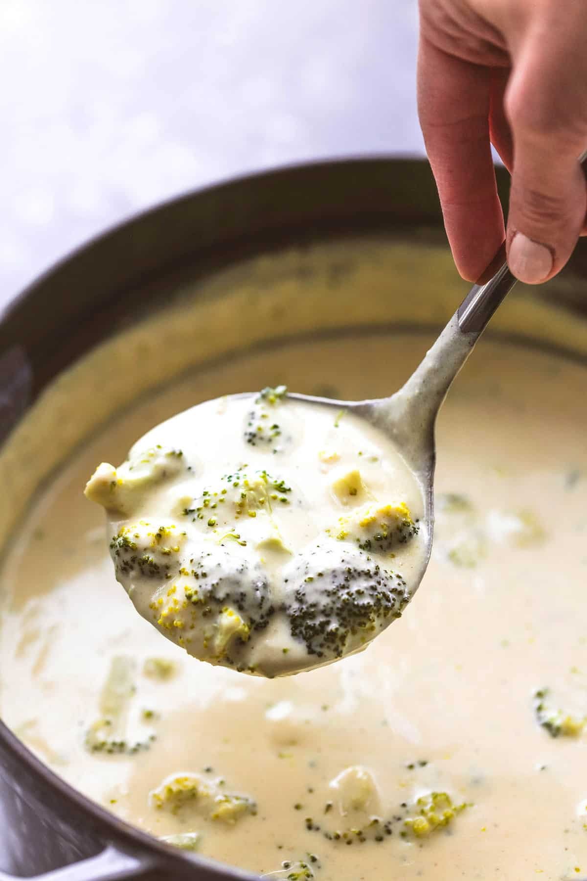 a hand lifting a serving spoon of broccoli cheese soup from a pot of soup.