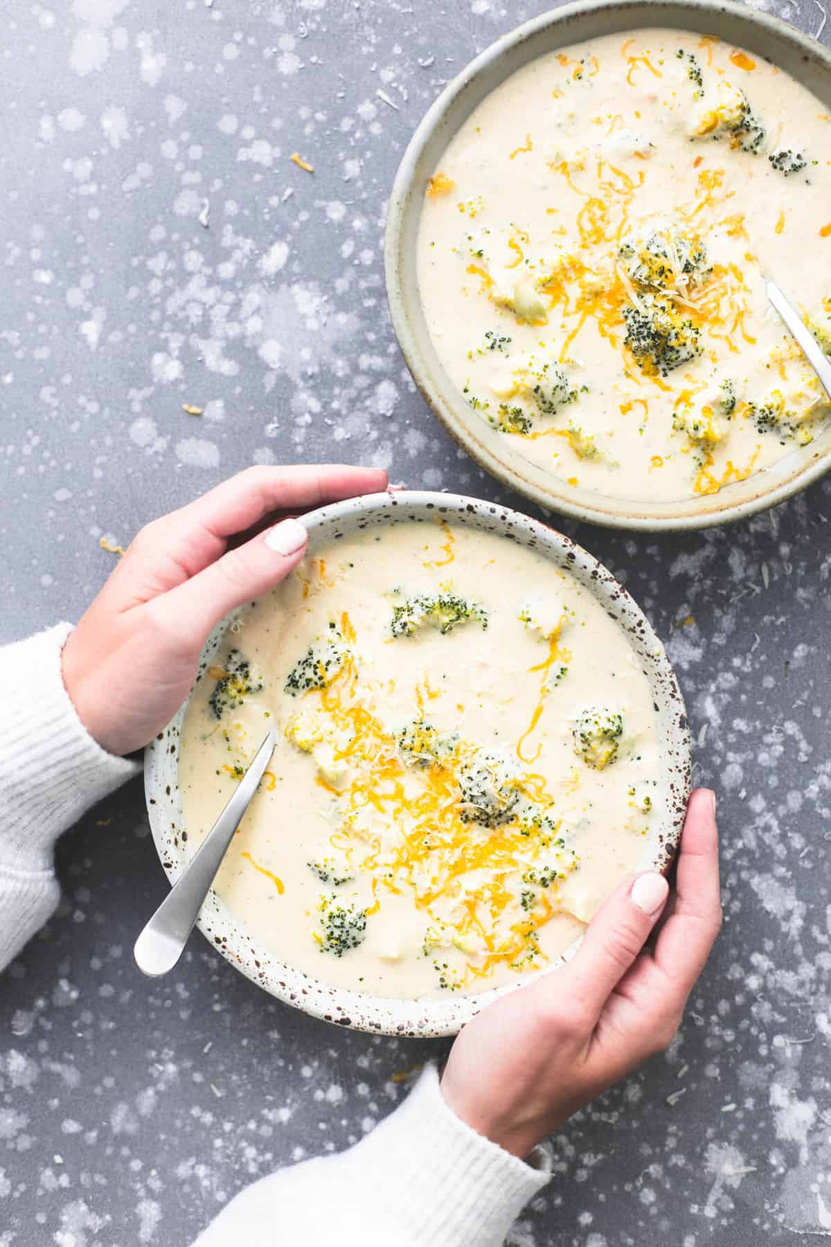 top view of a pair of hands holding a bowl of broccoli cheese soup with a spoon with another bowl of soup on the side.