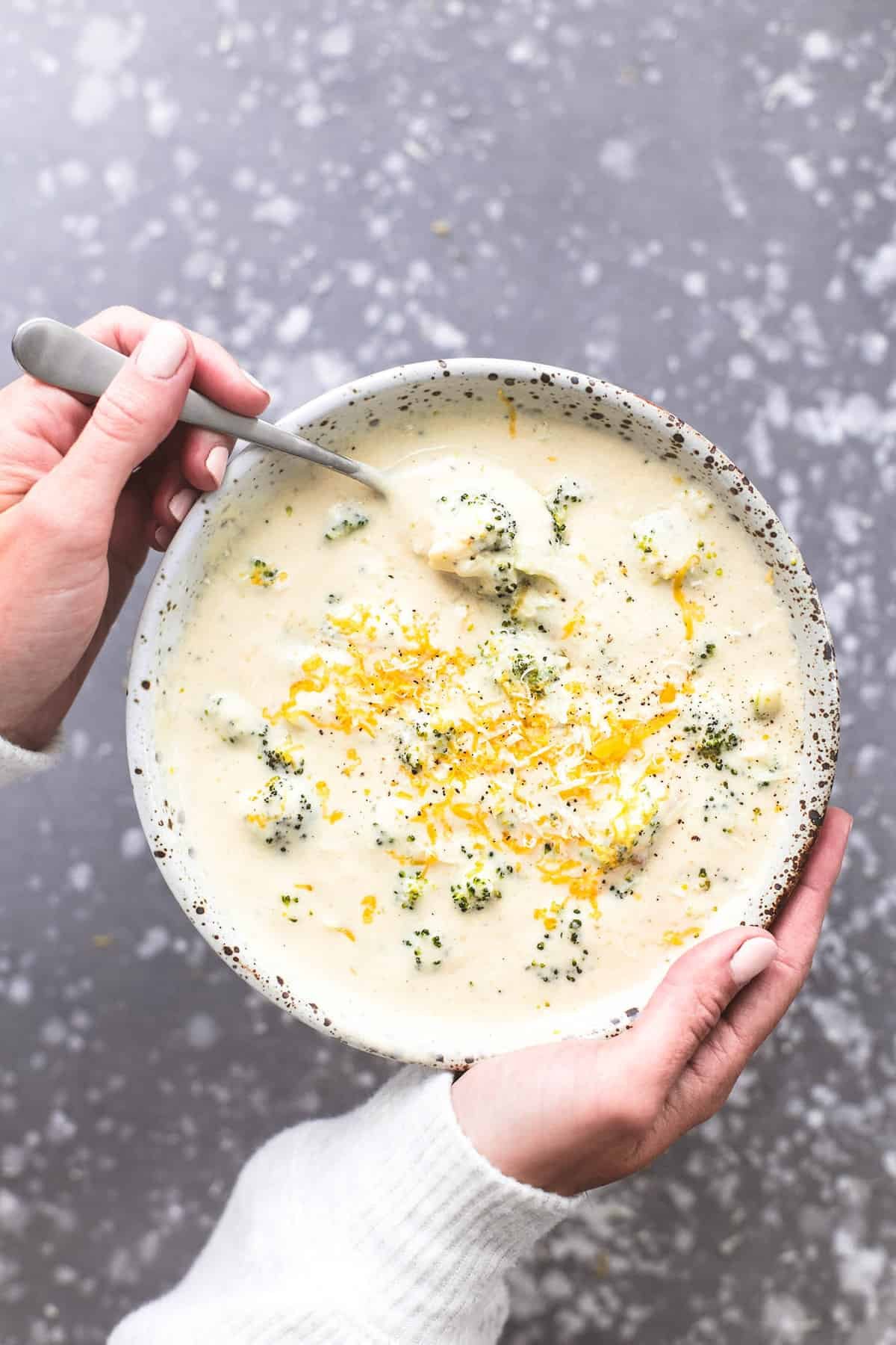 top view of a pair of hands holding a bowl of broccoli cheese soup with a spoon.