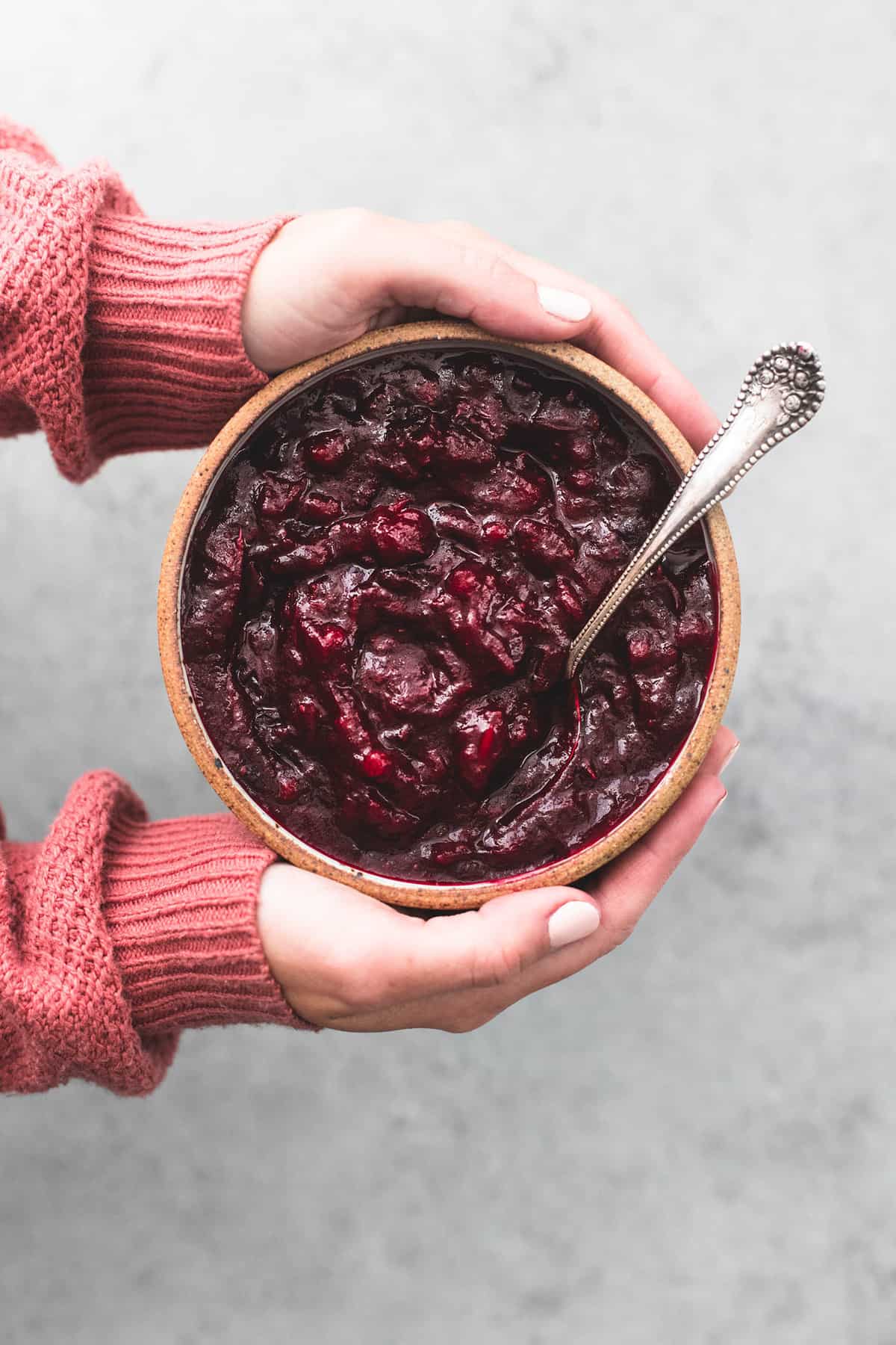 top view of a pair of hands holding a bowl with cranberry sauce and a spoon in it.