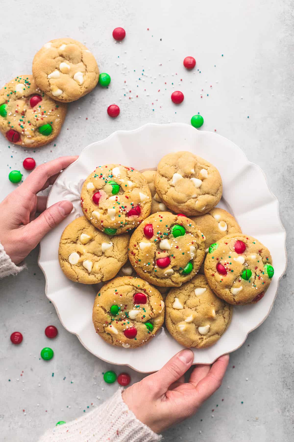 a pair of hands holding a plate of Santa's cookies (double chocolate M&M cookies) with M&Ms and more cookies on the side.