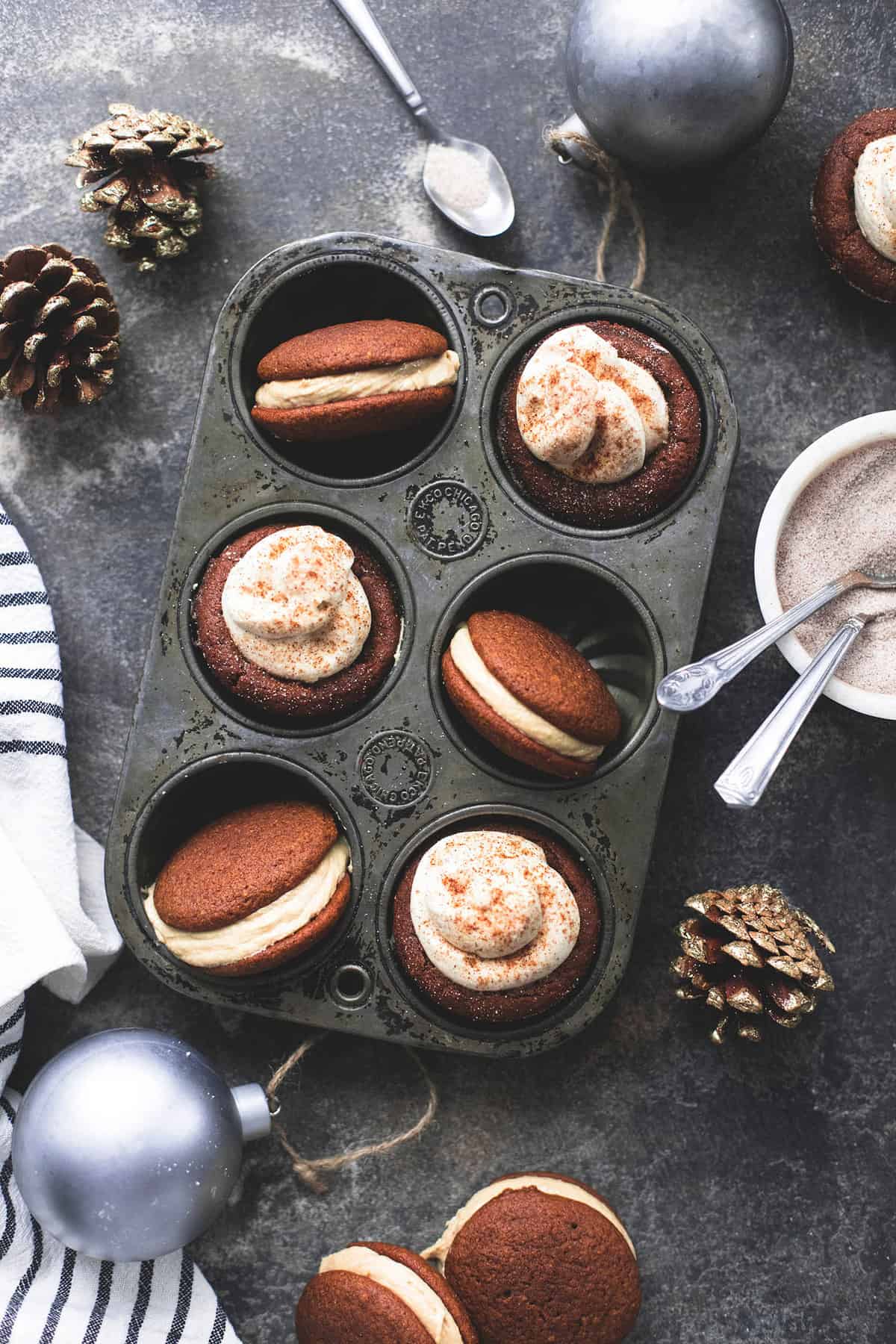 top view of gingerbread cheesecake cups and whoopie pies with caramel frosting in a muffin tin surrounded by pinecones, silver Christmas ornaments, and a bowl of cinnamon with spoons.