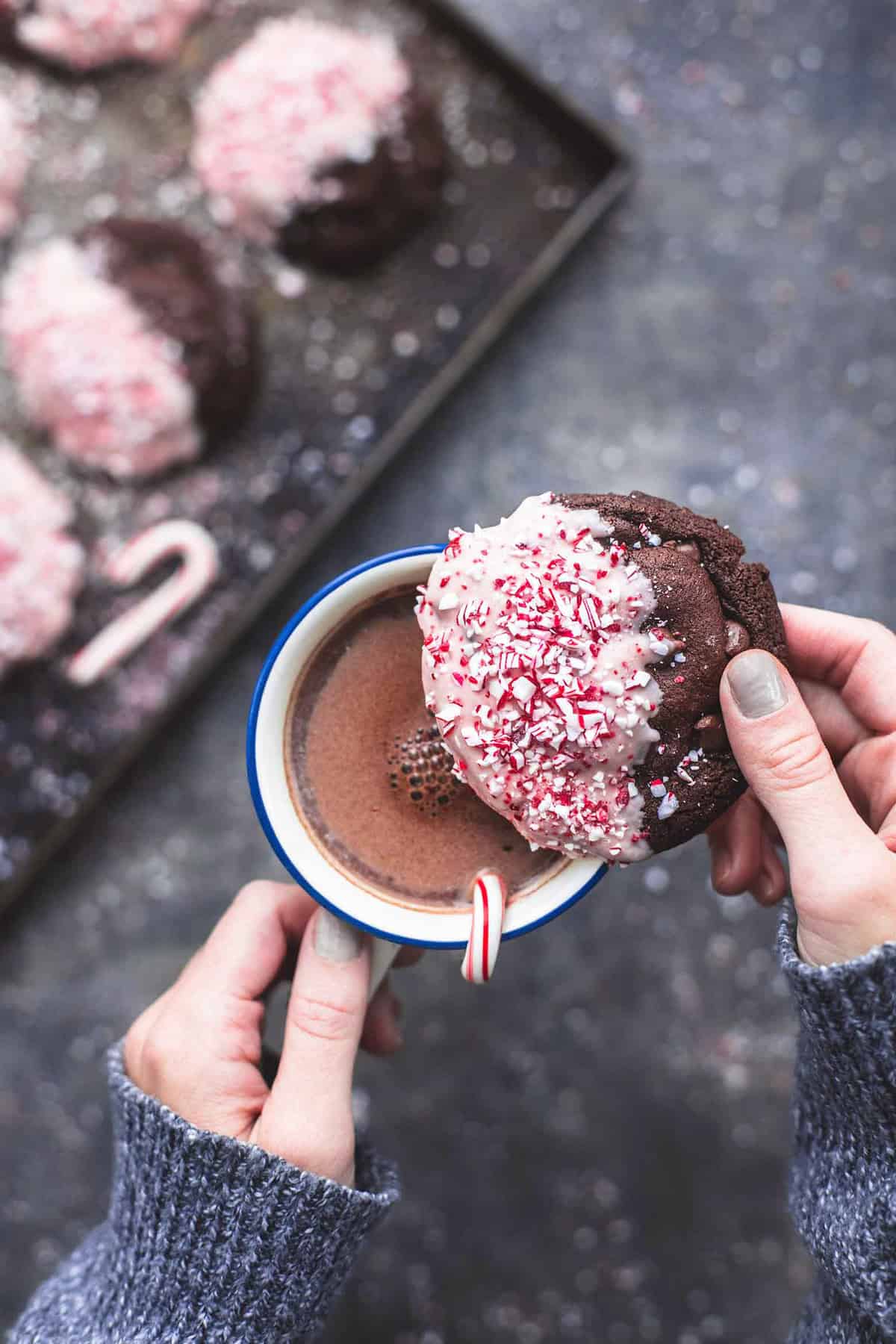 top view of a pair of hands with one hand holding the handle of a mug of hot chocolate and the other picking up a double chocolate peppermint bark cookie form on top of the mug being held above a pan with more cookies.