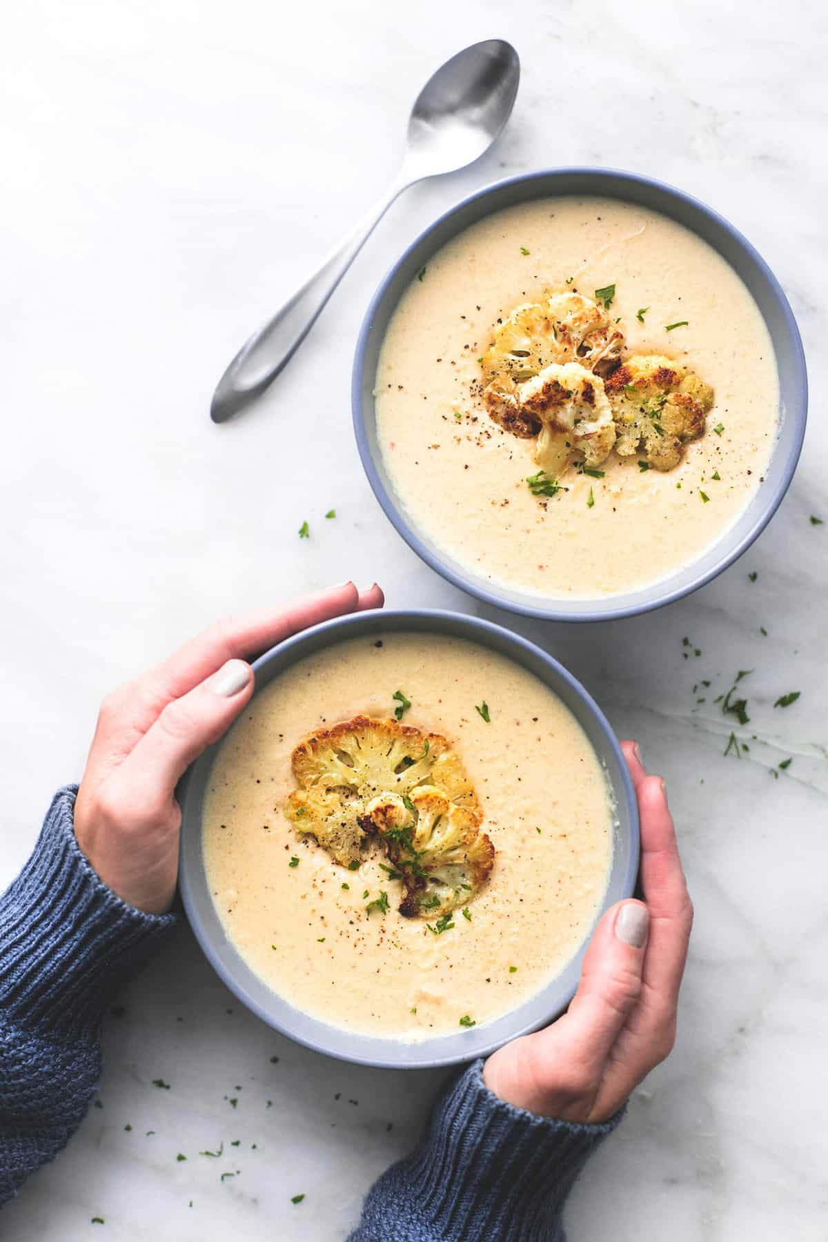 top view of a pair of hands holding a bowl of roasted cauliflower soup with a spoon and another bowl of soup on the side.