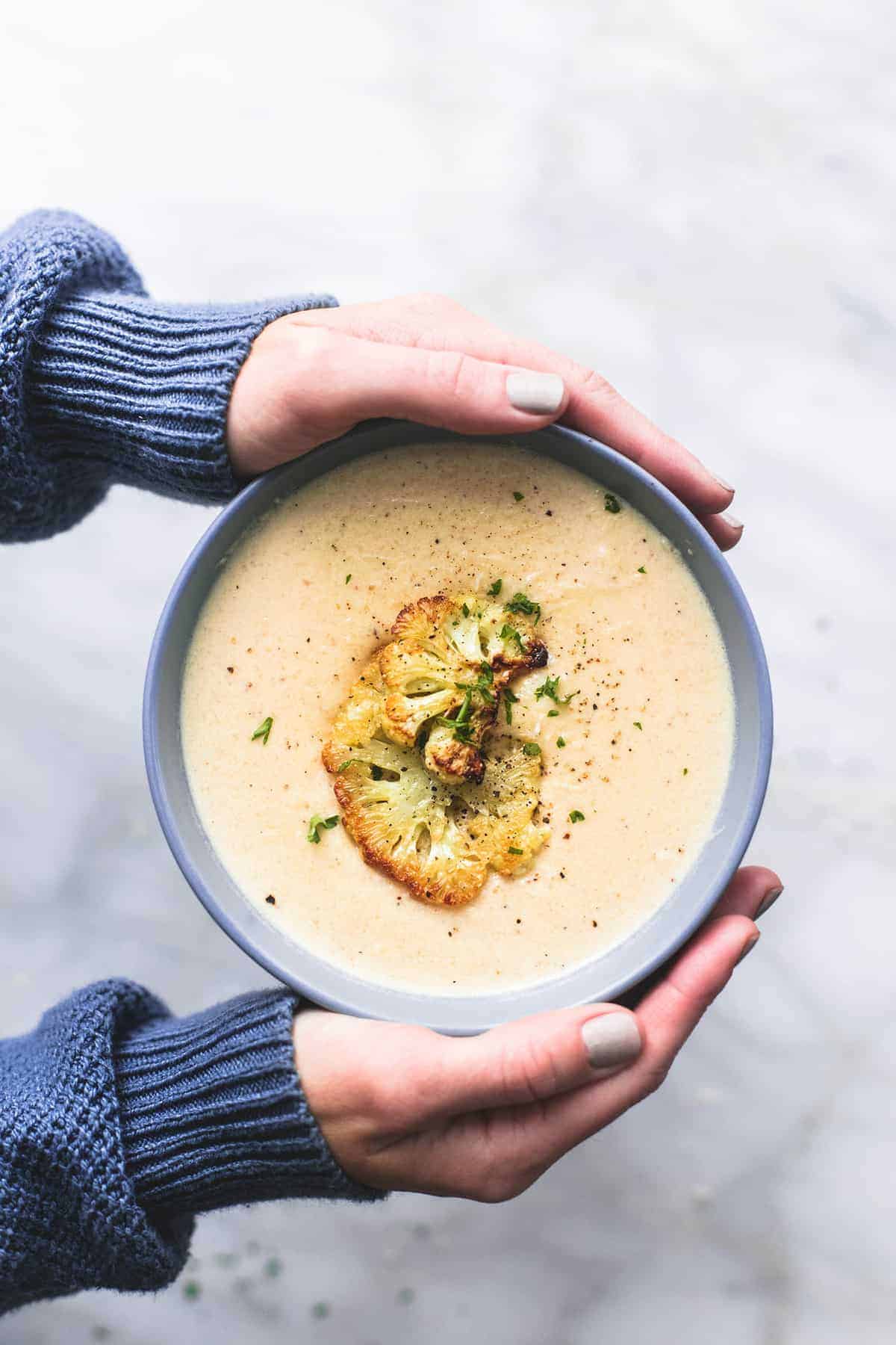 top view of a pair of hands holding a bowl of roasted cauliflower soup.