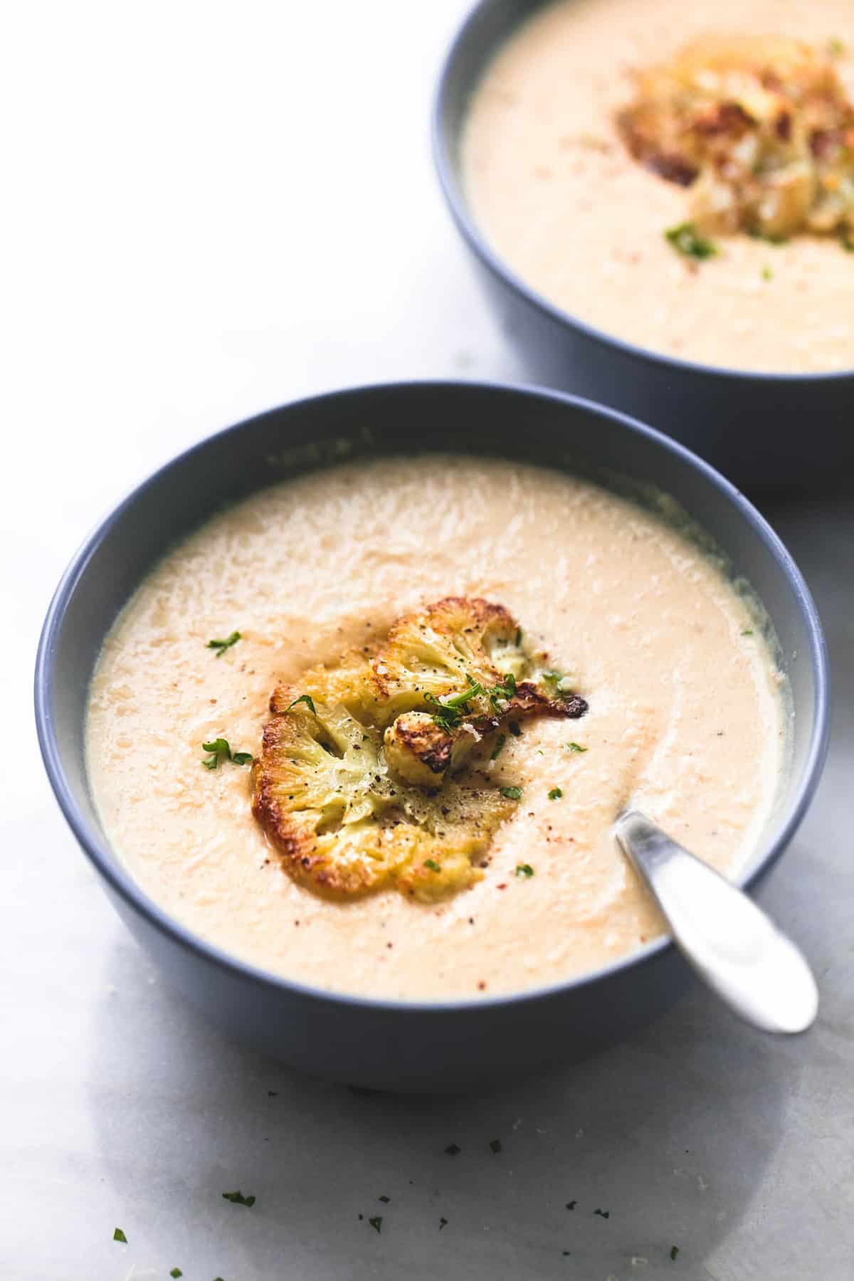 zuppa di cavolfiore arrosto con un cucchiaio in una ciotola con un'altra ciotola di zuppa in background.