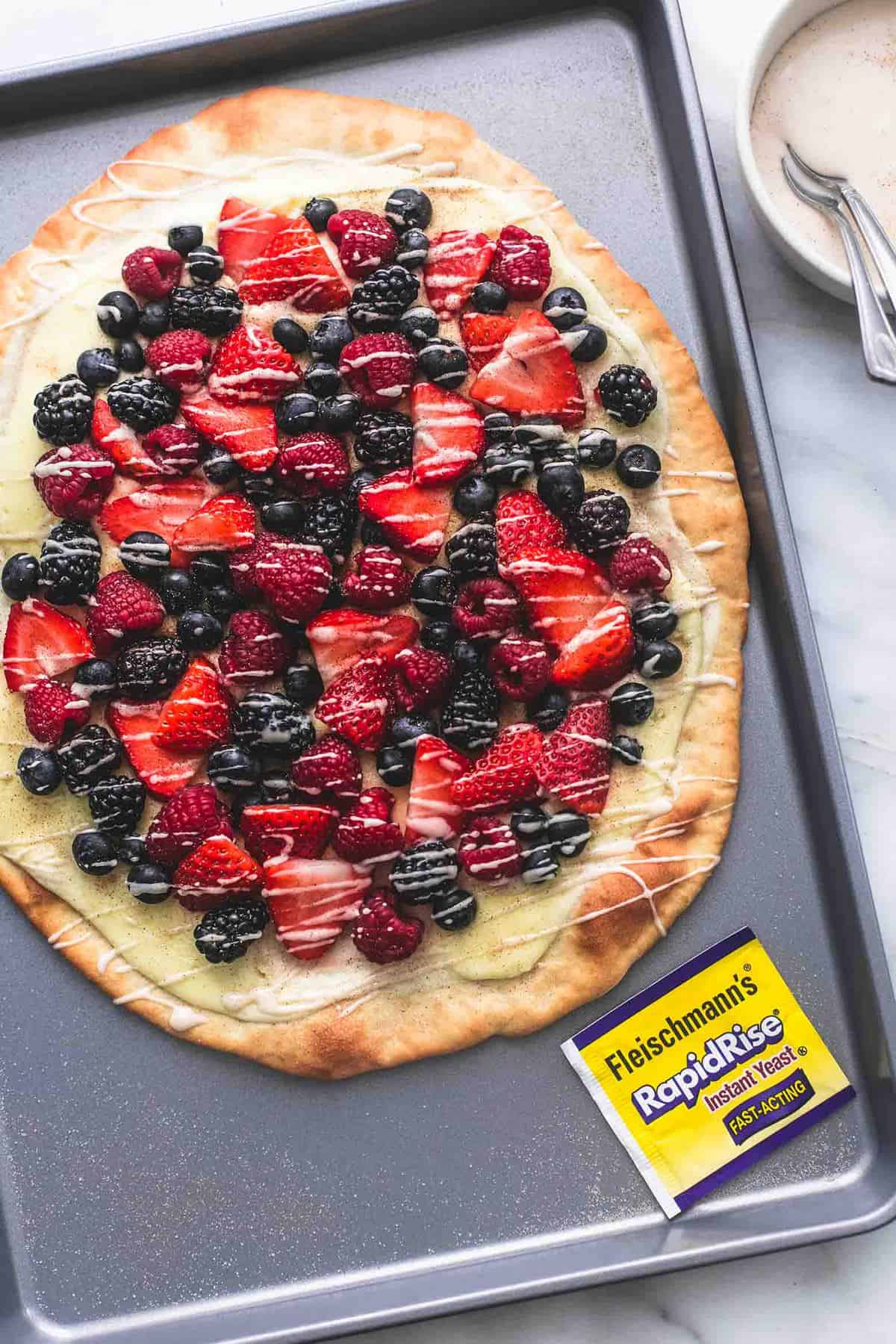 top view of mixed berry pizza with vanilla glaze and a package of RapidRise yeast on a baking sheet with a bowl of glaze on the side.