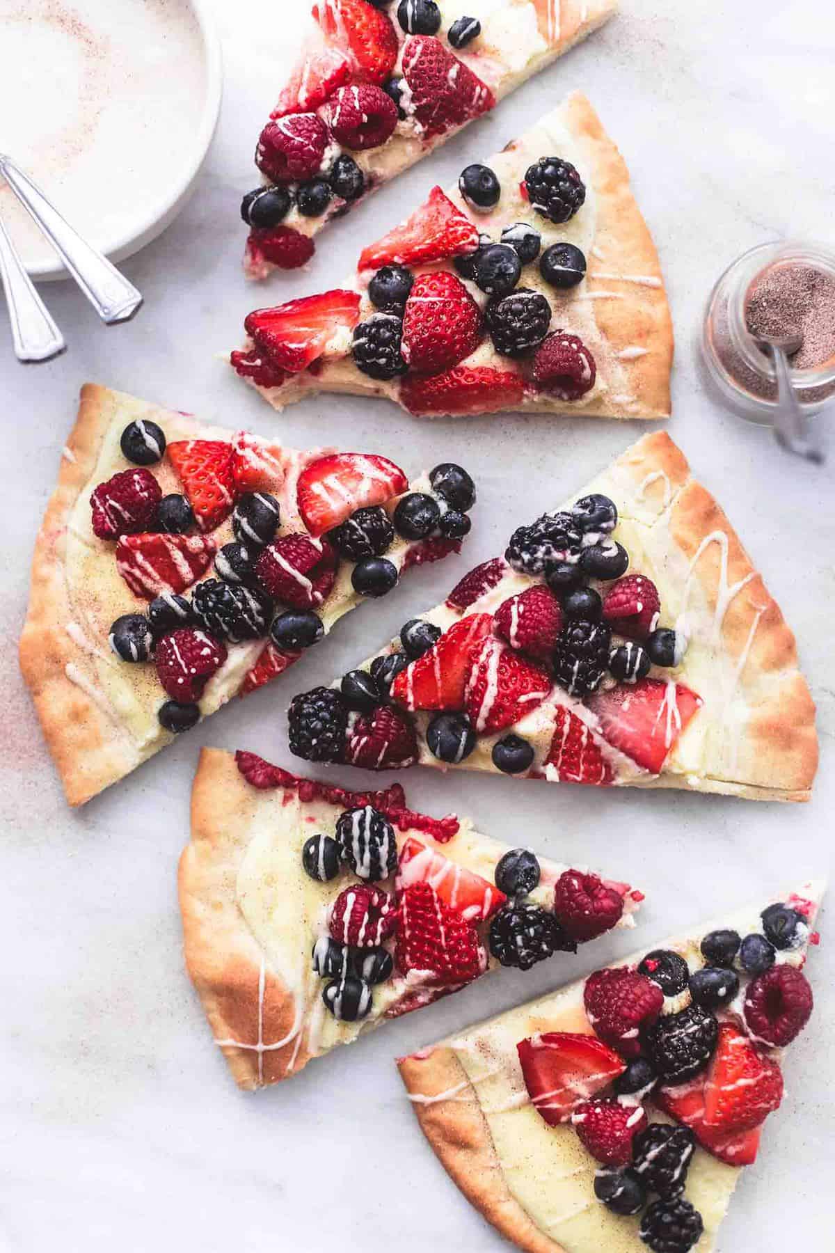 top view of mixed berry pizza with vanilla glaze slices with a jar of cinnamon sugar and a bowl of glaze on the side.