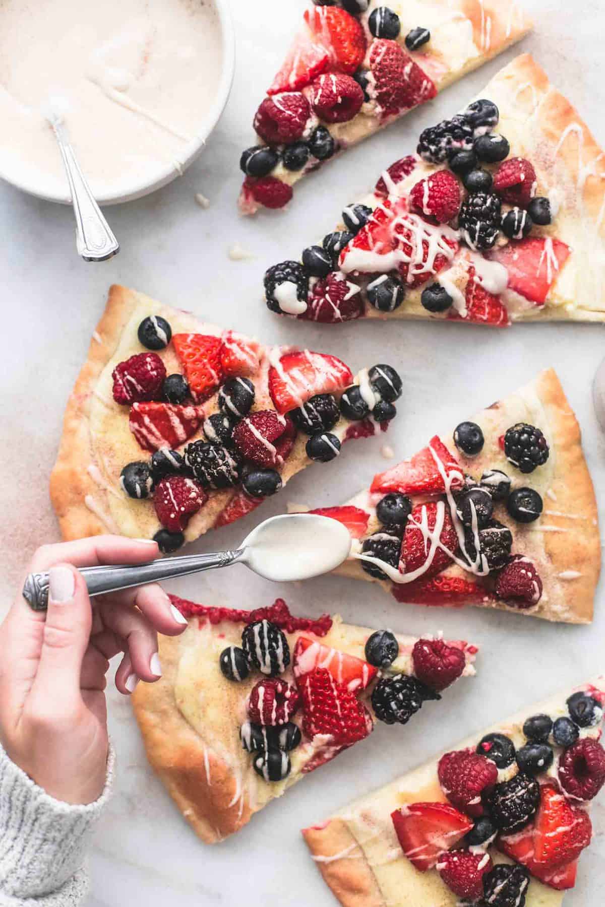 top view of mixed berry pizza with vanilla glaze slices with a bowl of glaze and a hand drizzling glaze over a slice with a spoon.