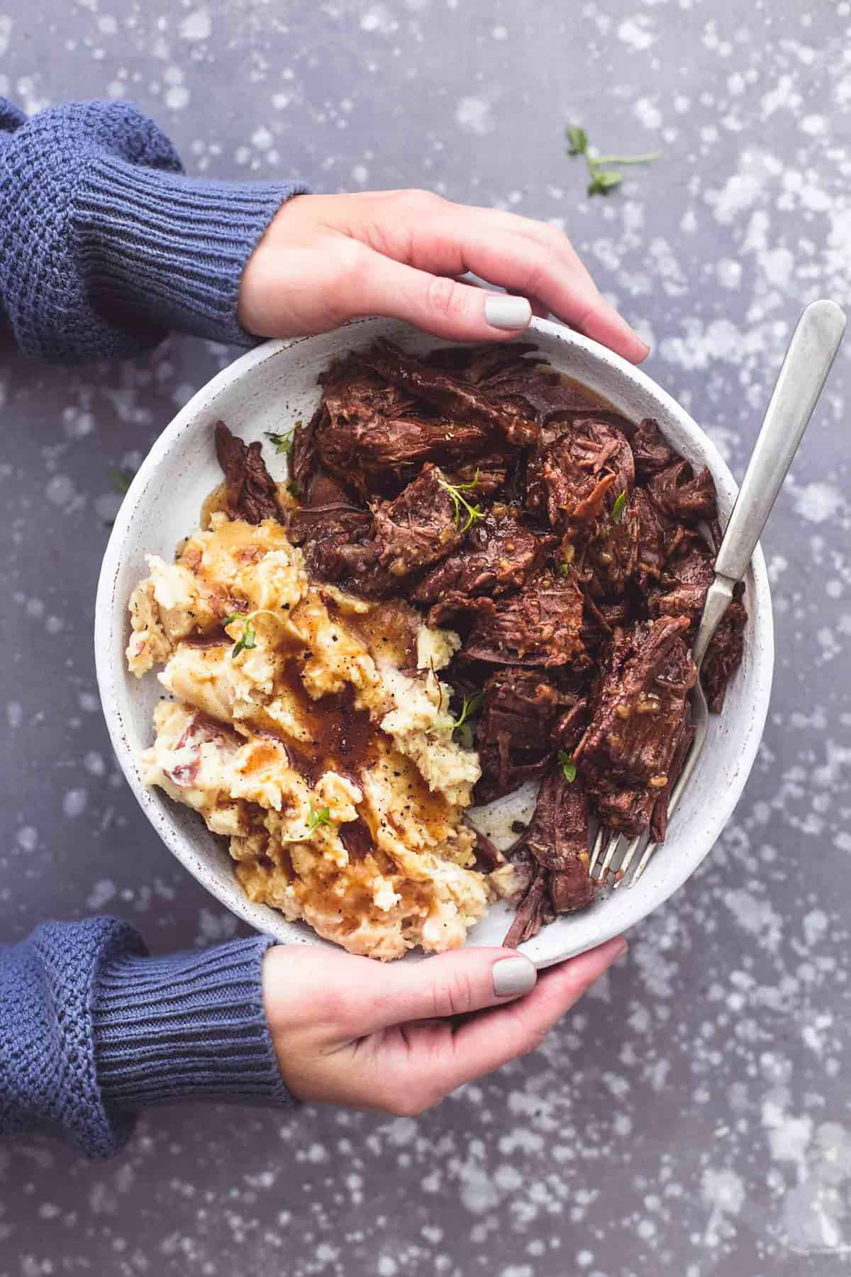 top view of a pair of hands holding a plate of instant pot pot roast with mashed potatoes and gravy with a fork.